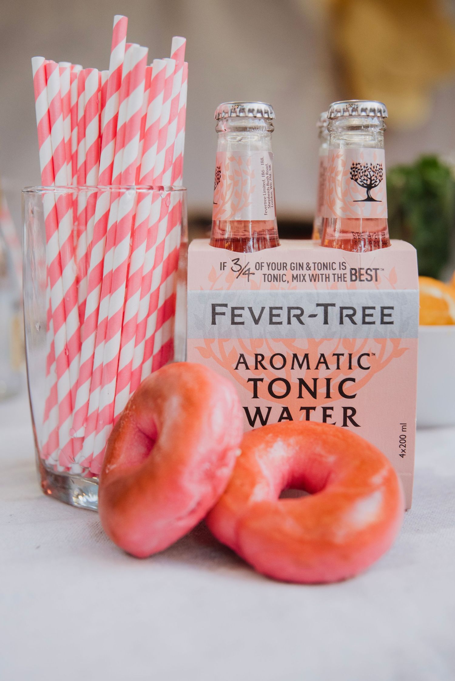 Pink glazed donuts beside Fever-Tree Aromatic Tonic Water bottles and pink striped paper straws in a glass.
