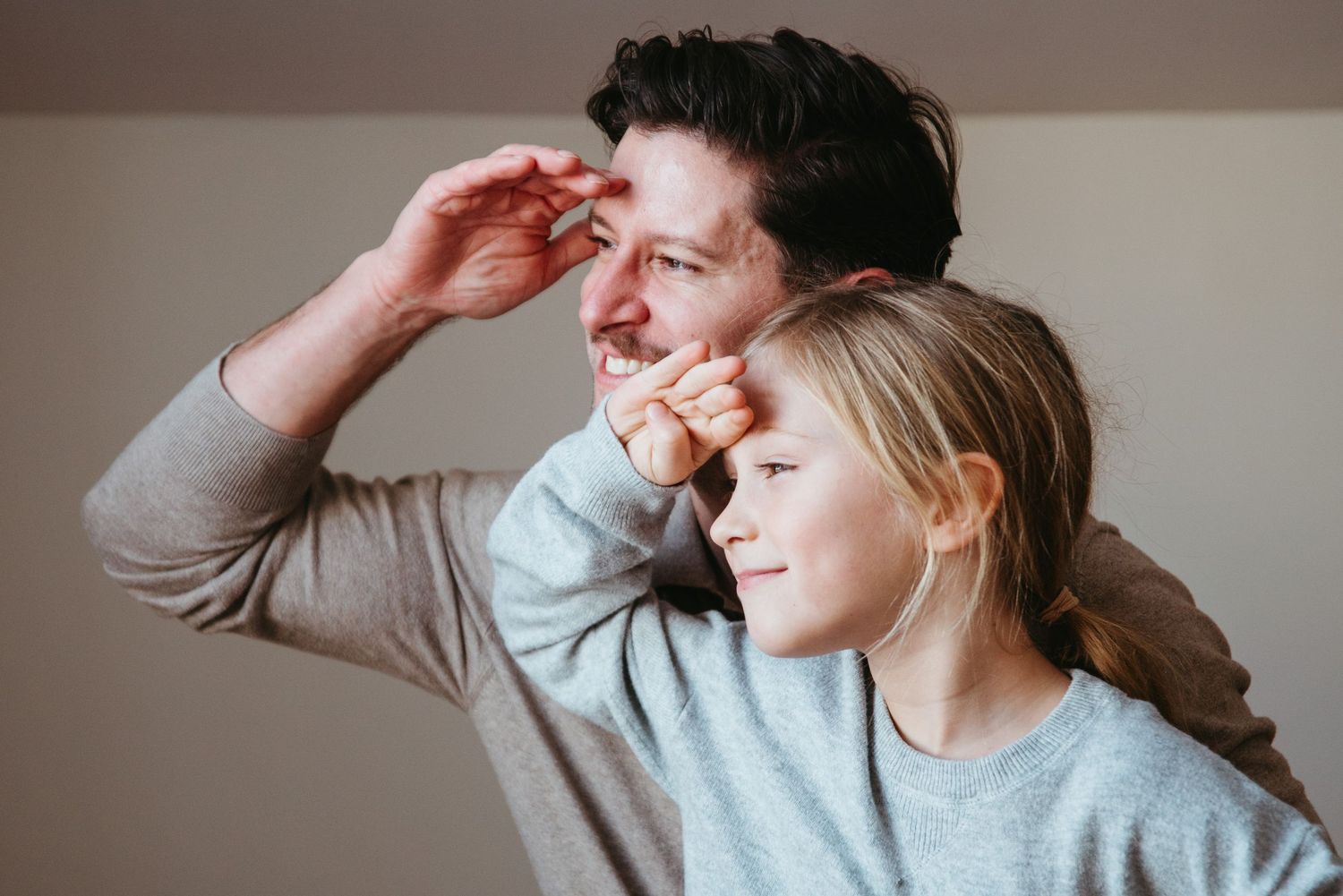 A father and young daughter in matching gray sweaters look upward together, sharing a joyful moment of curiosity.
