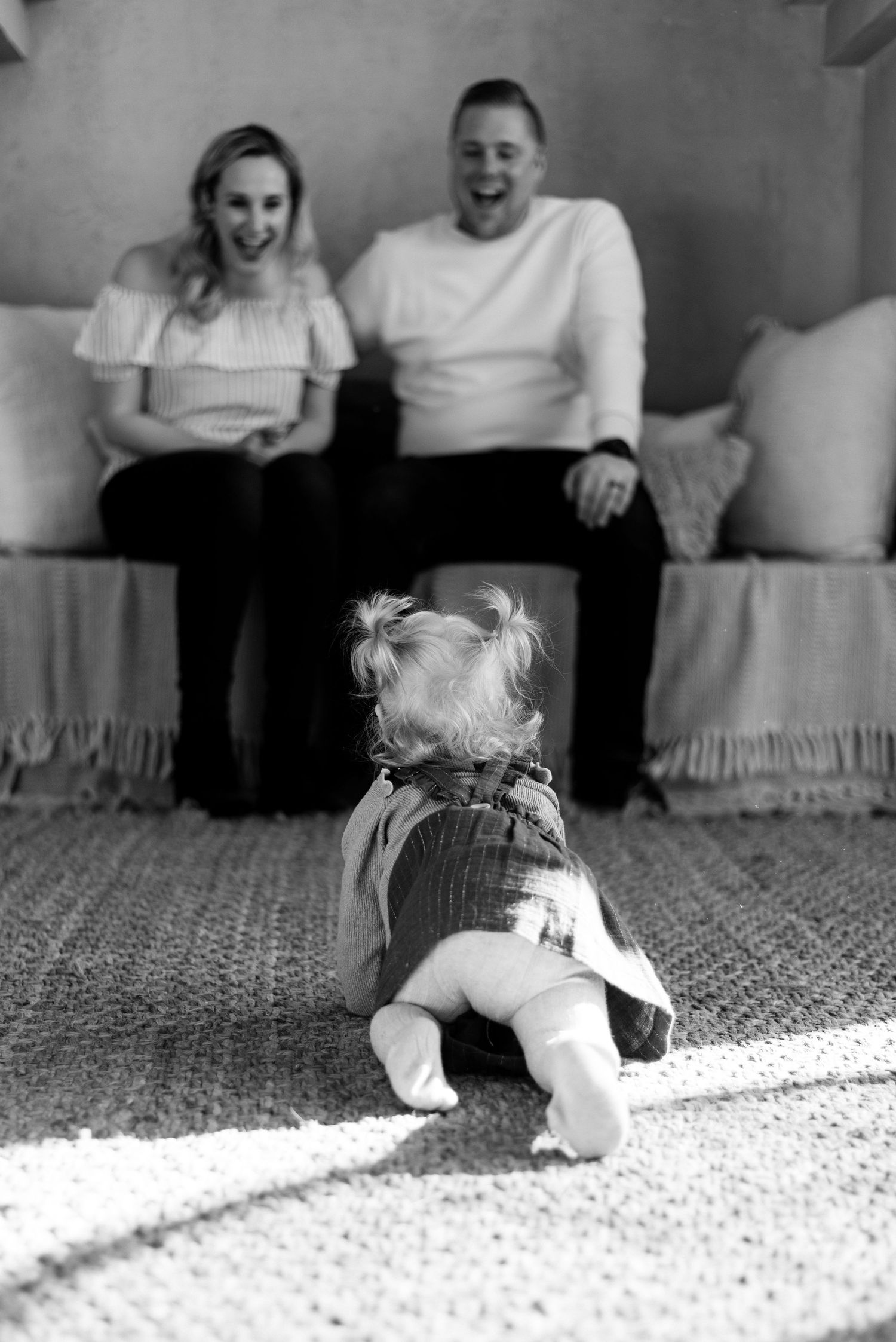 Black and white photo of a baby sitting on a rug facing laughing parents seated on a couch.