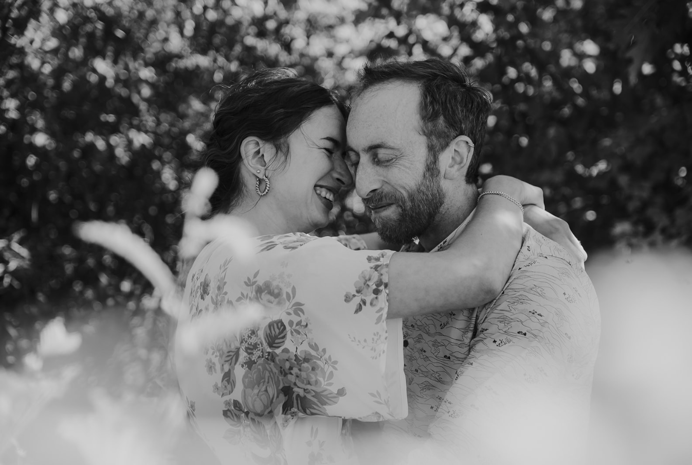 Black and white photo of a joyful couple embracing and laughing together outdoors among lush greenery.