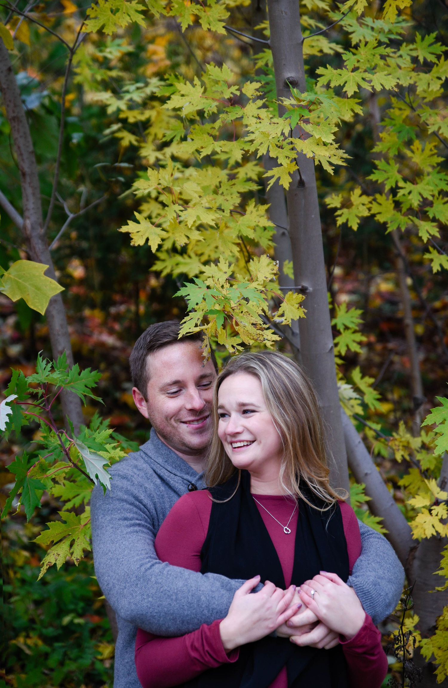 Couple embracing outdoors among vibrant autumn foliage, woman smiling in burgundy top, man in gray sweater hugging her from behind.