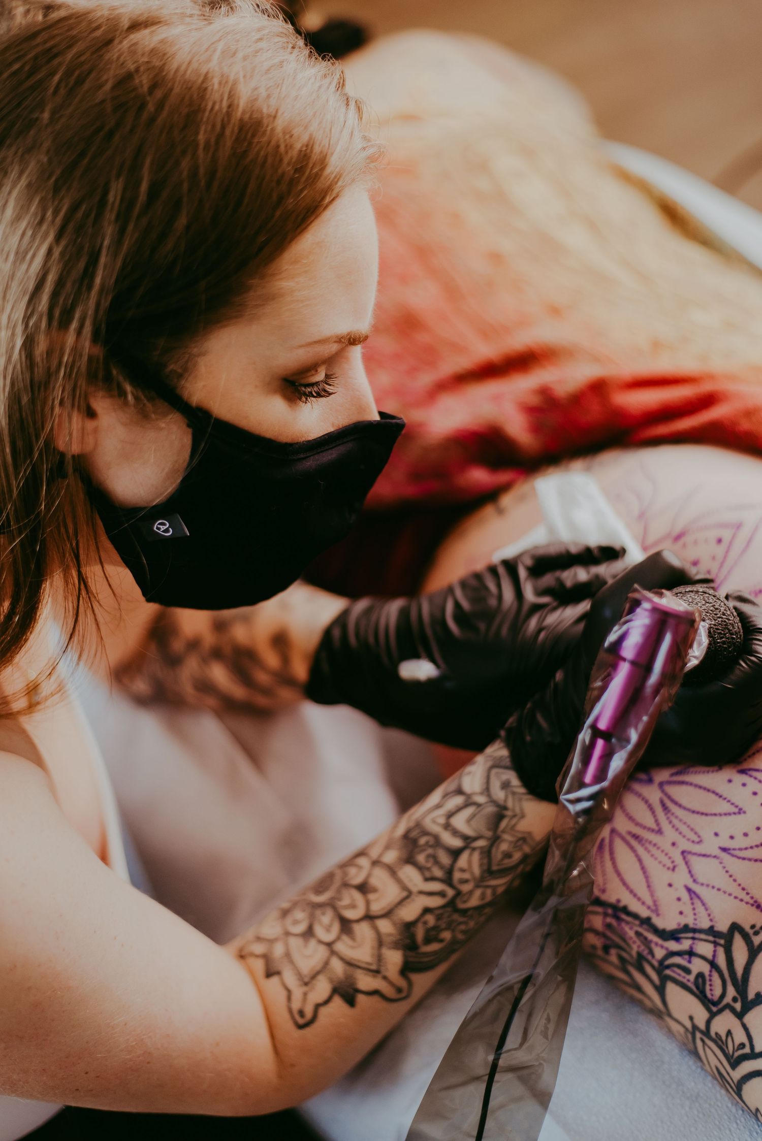 Female tattoo artist wearing a black mask and gloves carefully tattooing an intricate design on a client.