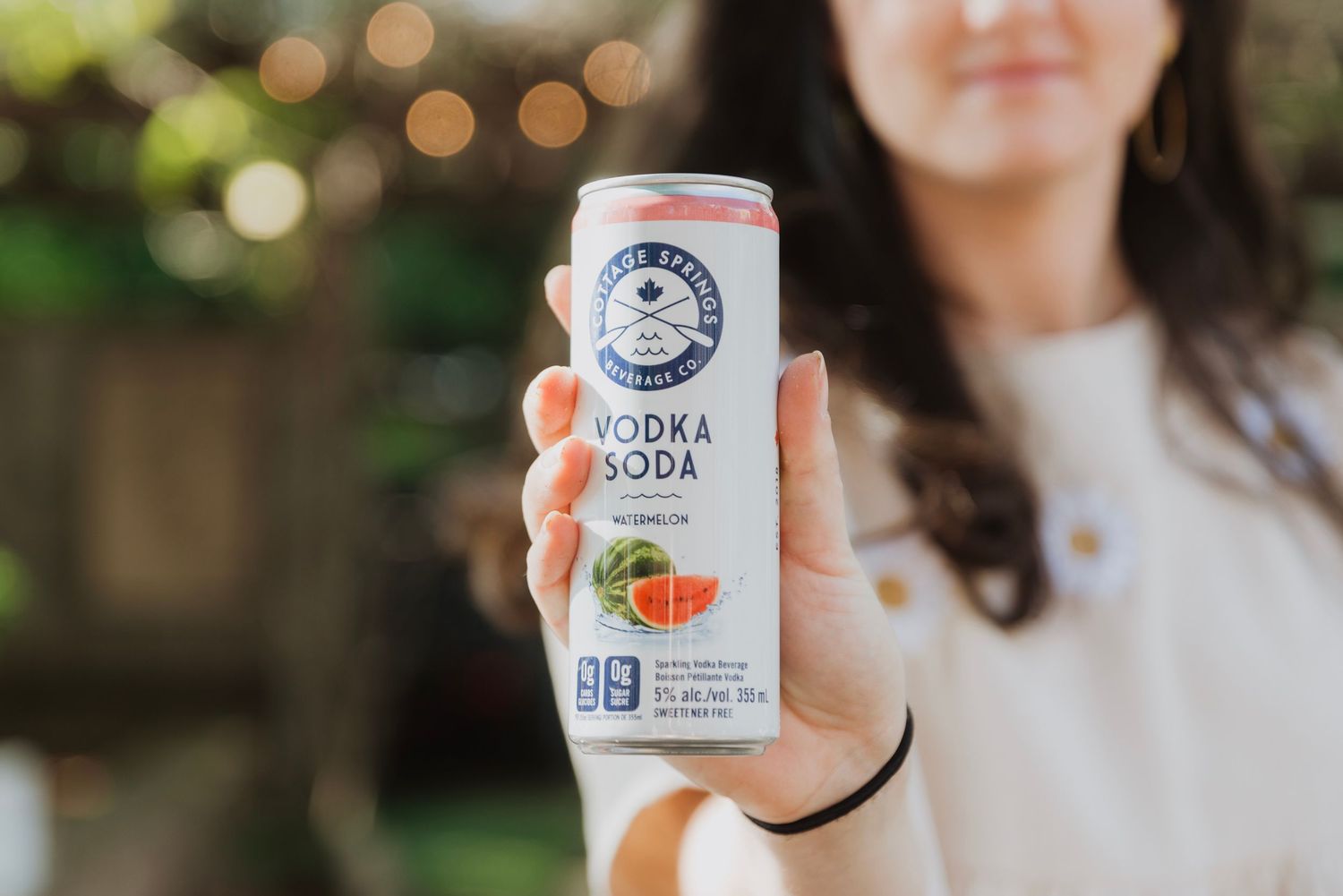 Woman holding a White Claw Watermelon Vodka Soda can outdoors with bokeh lights in background.