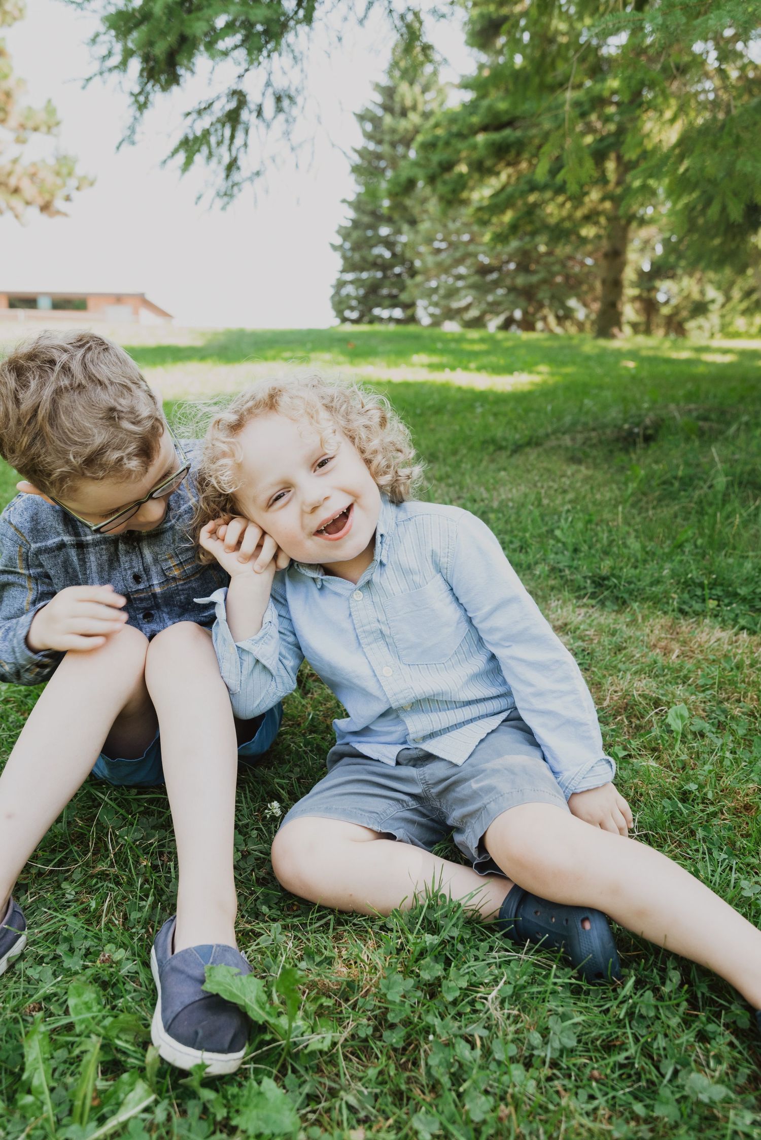 Two young boys sitting on green grass in a park, laughing and whispering to each other on a sunny day.