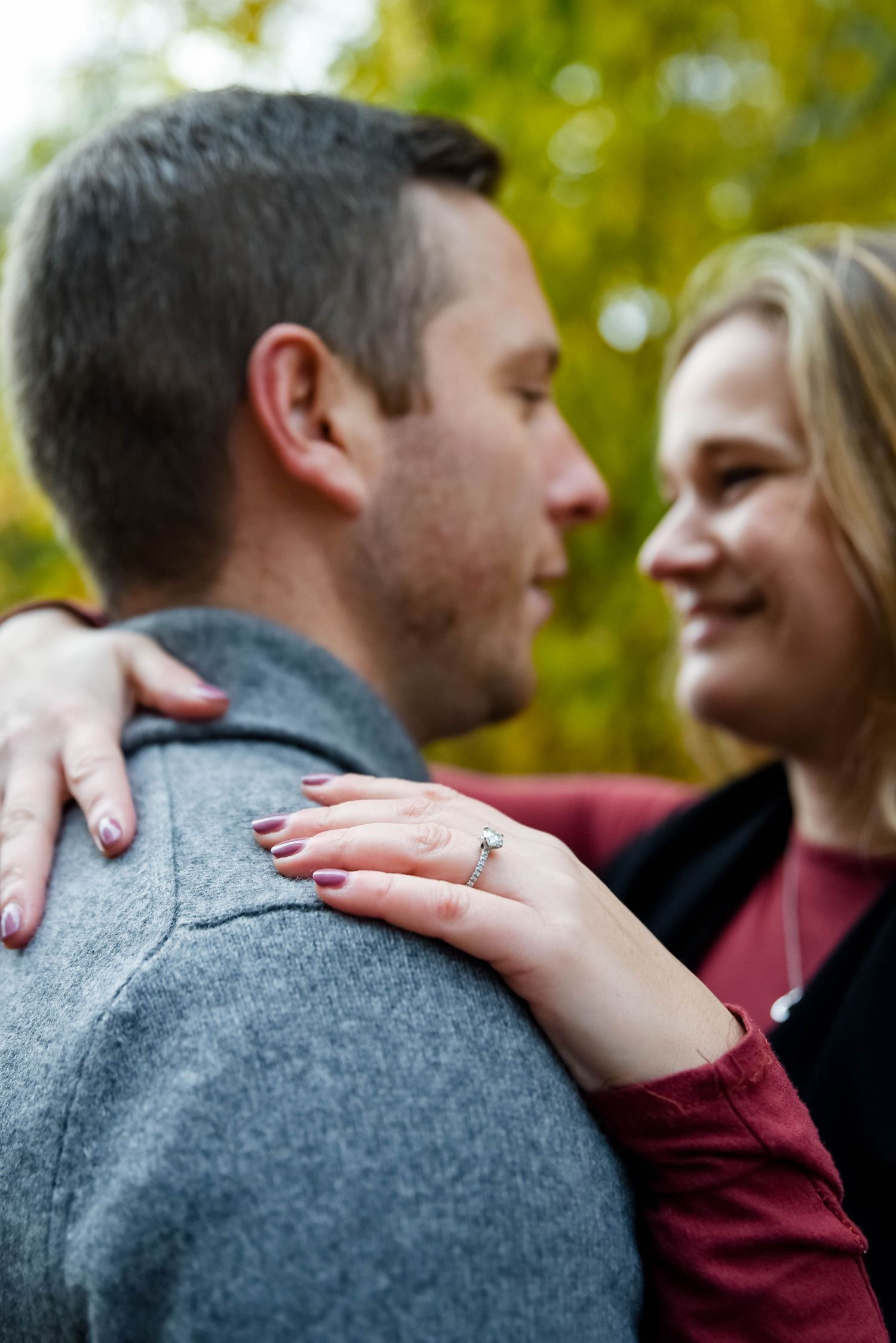Couple embracing outdoors in fall, woman smiling and showing engagement ring on man's shoulder.