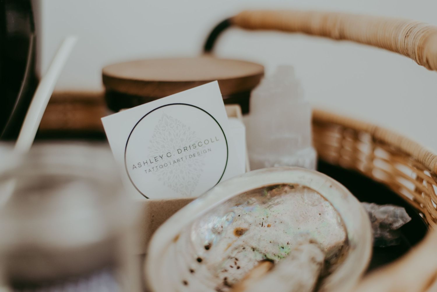 Wicker basket with Ashley & Driucci business card, wooden lid candle, and artisan soap on a white surface.