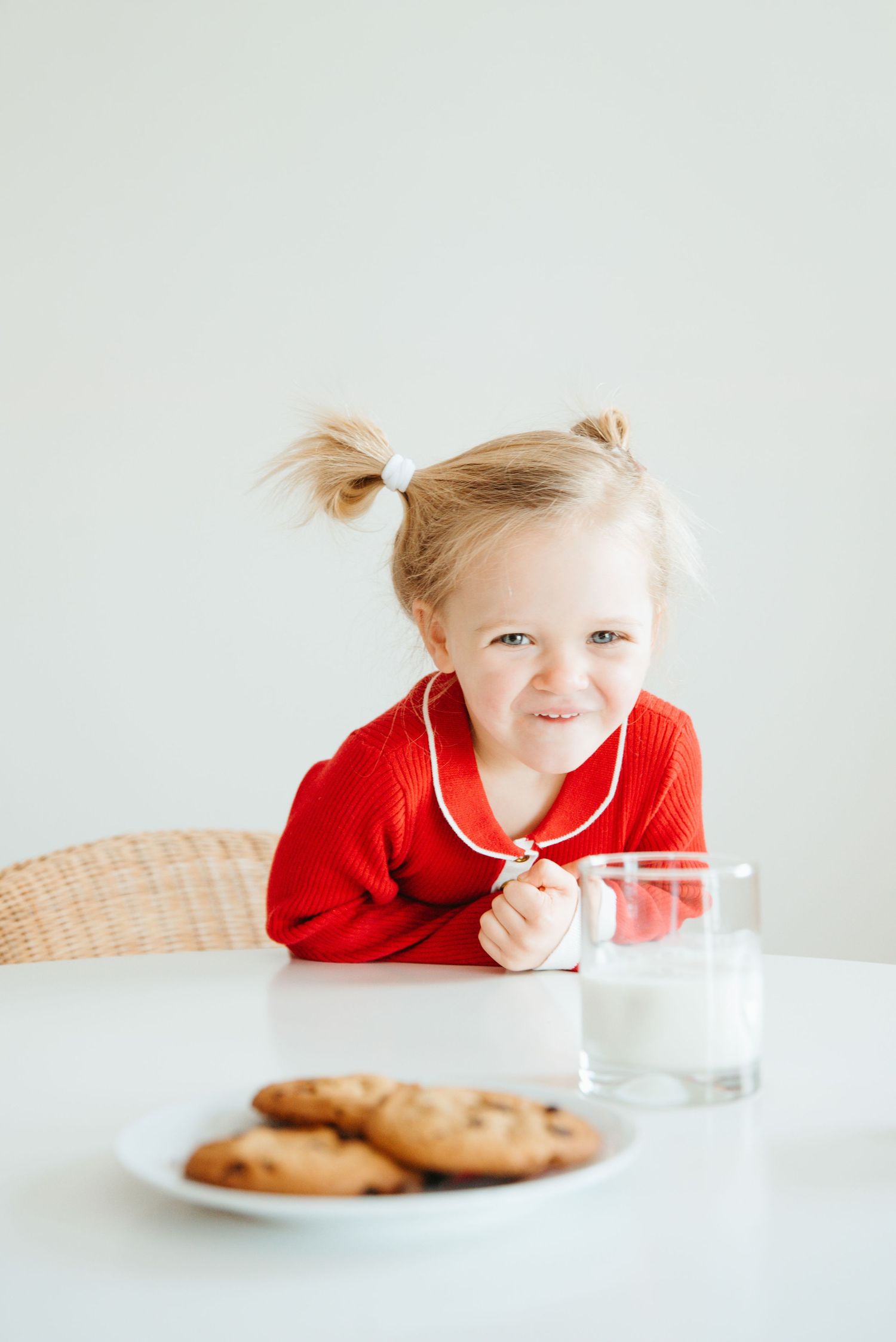 Smiling toddler girl with pigtails in red sweater sitting at white table with chocolate chip cookies and glass of milk.