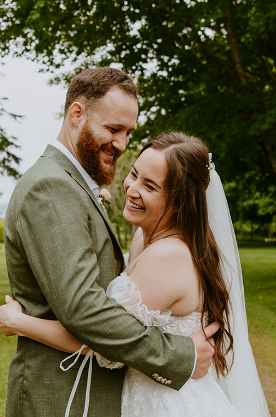 A smiling bride in a lace off-shoulder gown and veil embraces a bearded groom in a green suit outdoors.