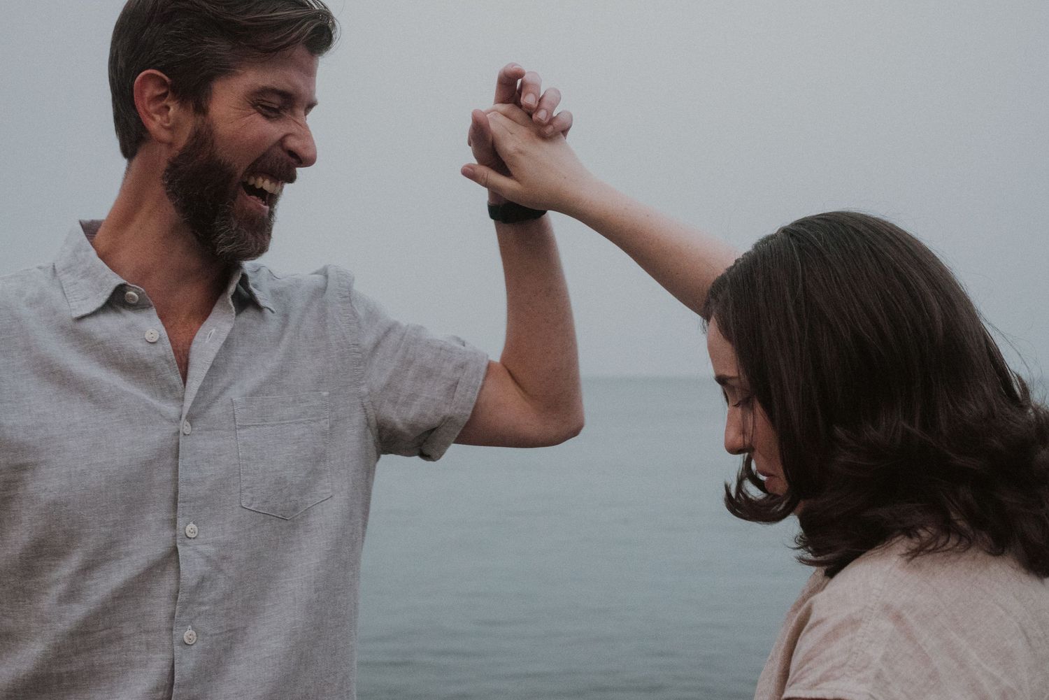 A happy couple dancing together near the ocean, the man laughing as he twirls the woman under his raised hand.