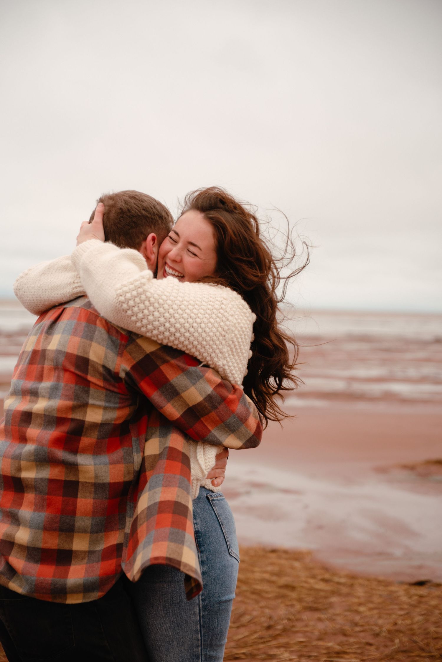 A couple shares a joyful embrace on a windy beach, the woman smiling warmly in a cream sweater and jeans.