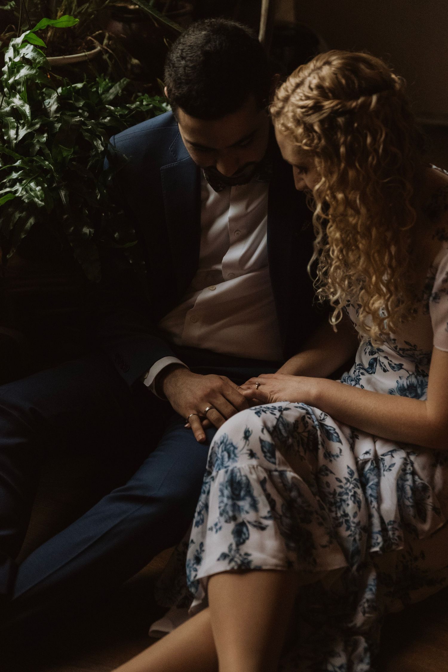 Couple sharing an intimate engagement moment, woman in floral dress with curly hair, man in navy suit, holding hands indoors.