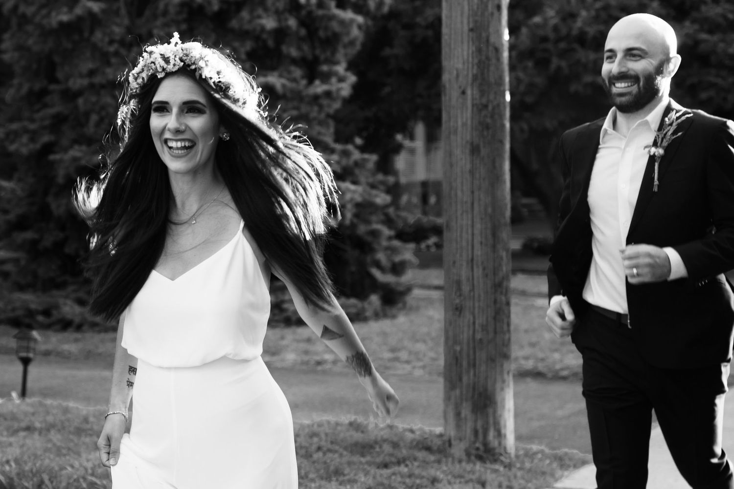 Black and white photo of a smiling bride in white dress with floral crown and groom in suit running outdoors.