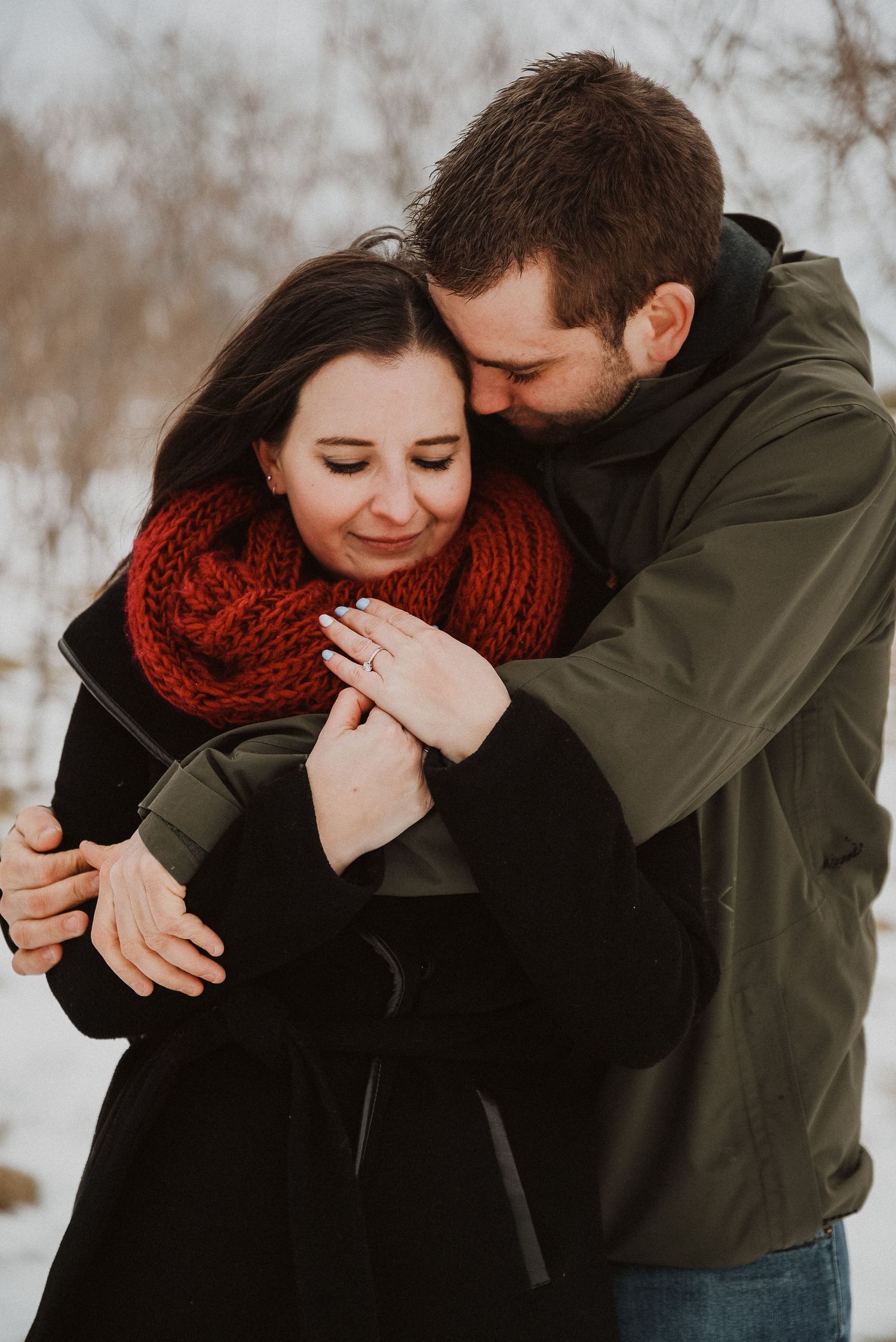 Couple embracing in snowy winter setting, woman in black coat and red scarf, man in olive jacket nuzzling her cheek.
