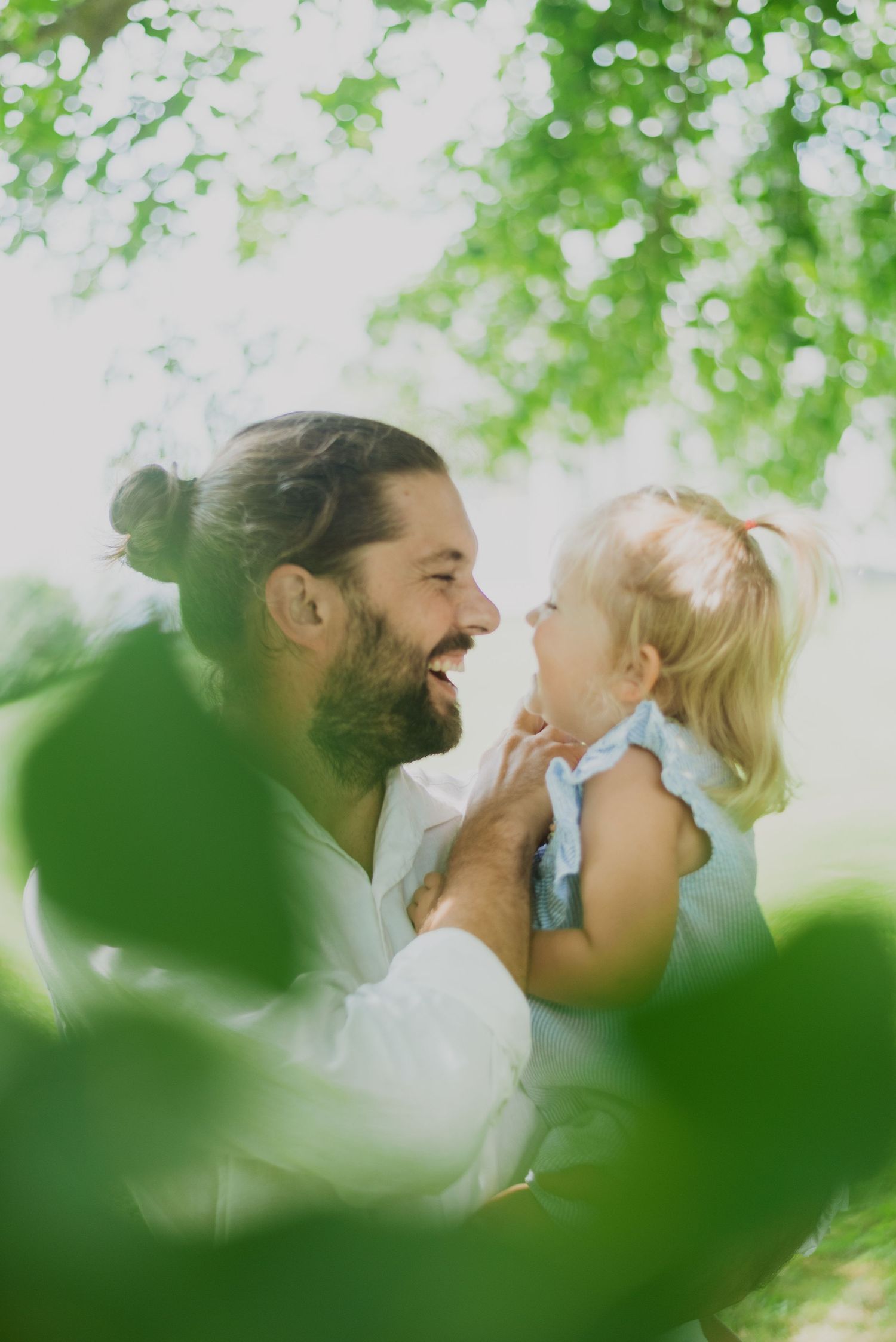 Bearded man with bun hairstyle smiling and holding a blonde toddler girl outdoors among green trees.