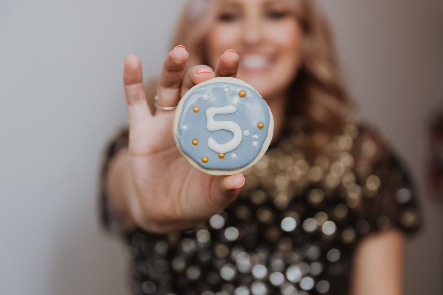 Woman in sequin dress holding a blue frosted cookie decorated with number 5 and gold sprinkles.