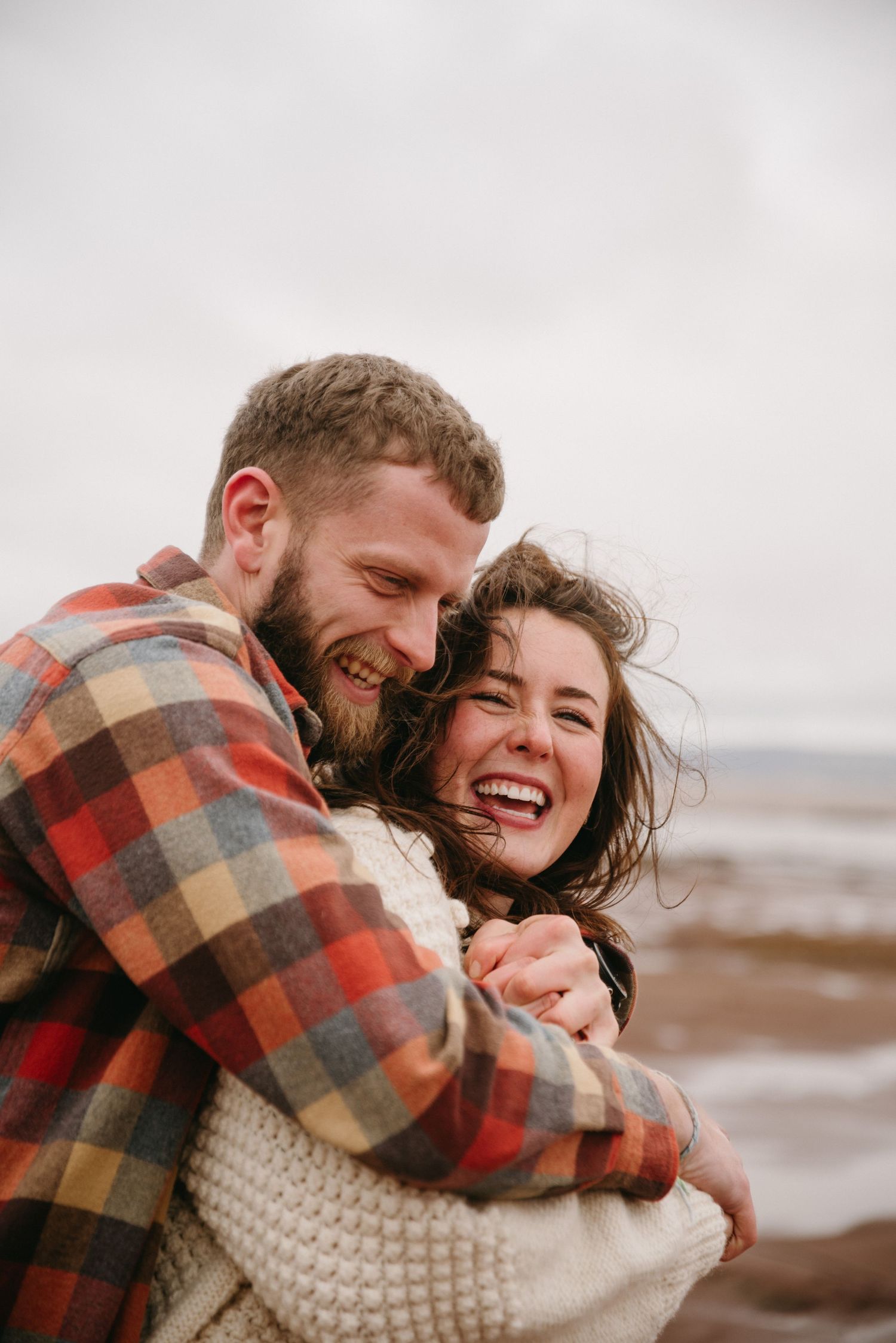 A couple shares joyful laughter and embraces on a cloudy beach while wearing cozy sweaters and plaid.