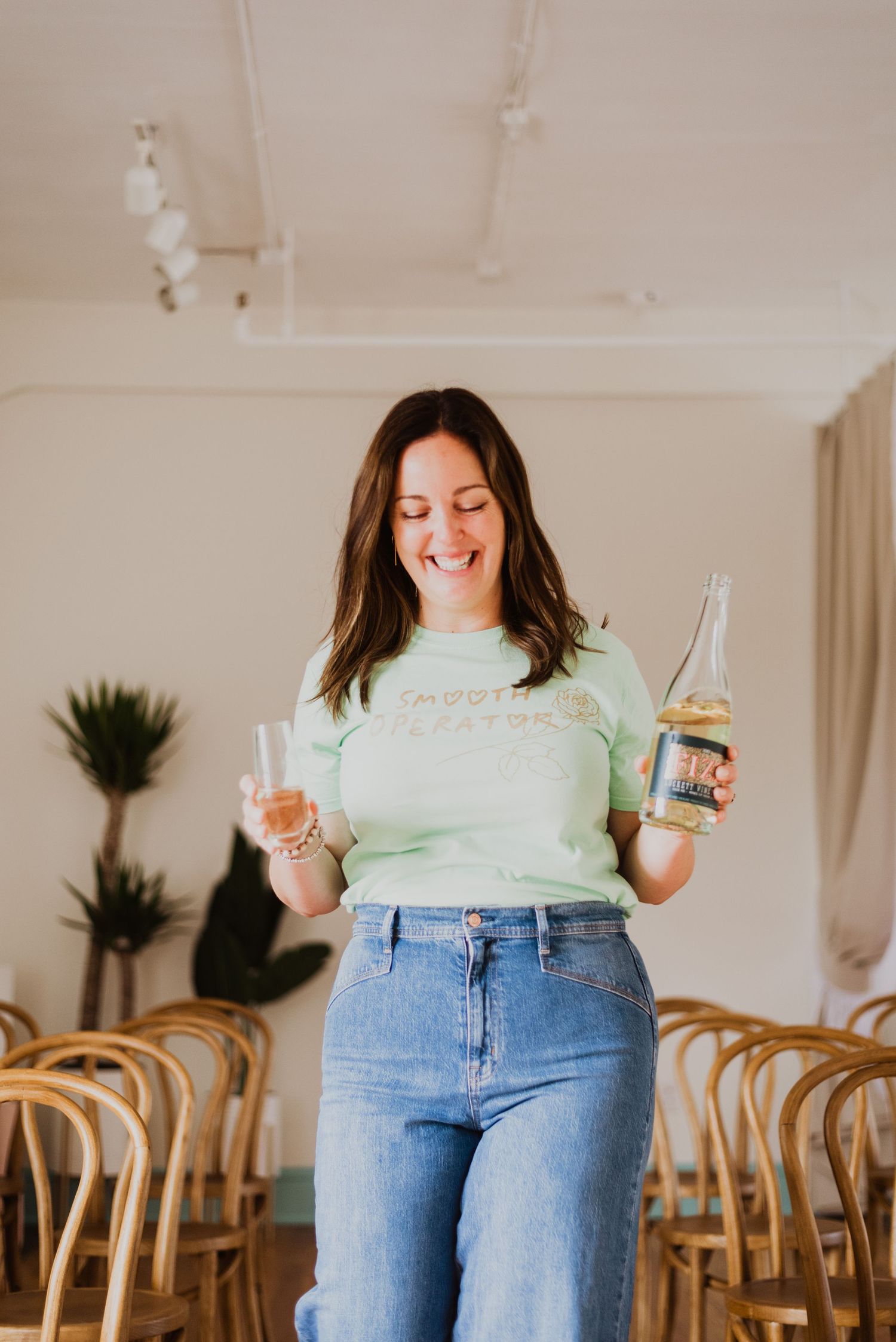 Smiling woman in mint green tee and jeans holding a wine bottle and glass indoors.