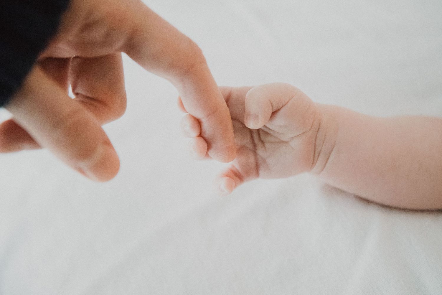 A baby gently grasping an adult's finger on a soft white background, symbolizing love and connection.