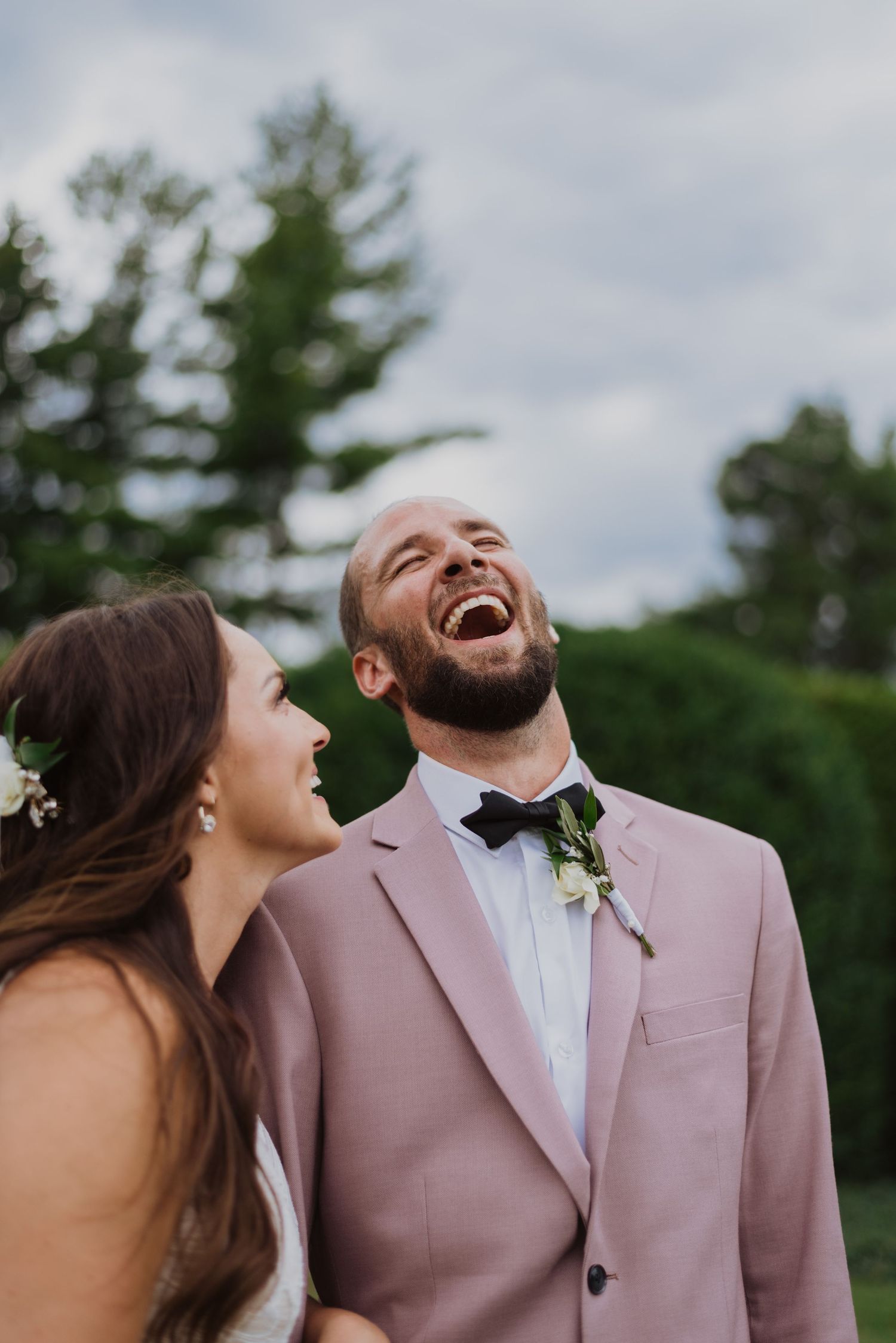 Joyful groom in pink suit laughing with bride during outdoor wedding ceremony amid lush green trees.