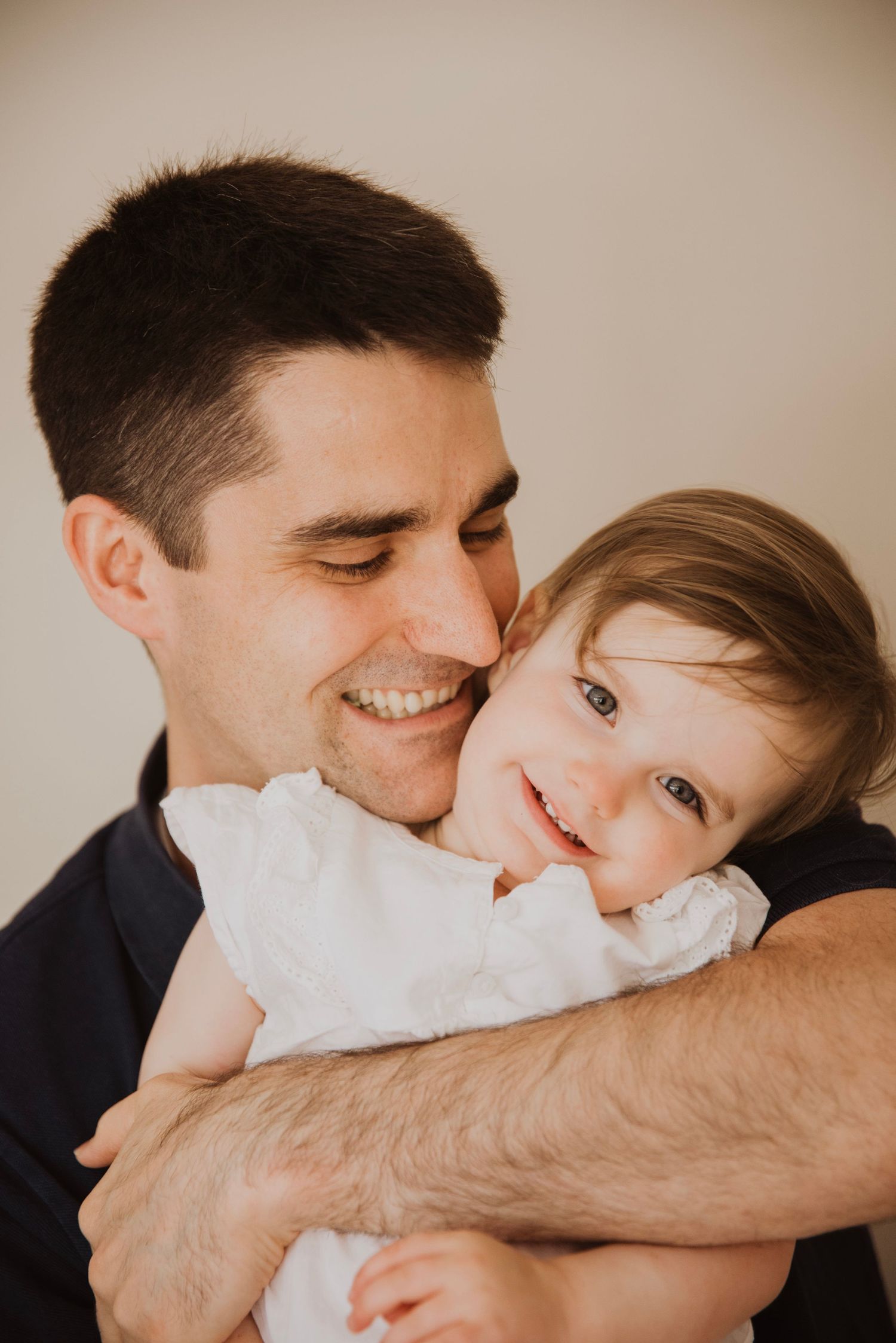 A smiling man tenderly holds a happy toddler girl in a white dress in a warm, candid family portrait.