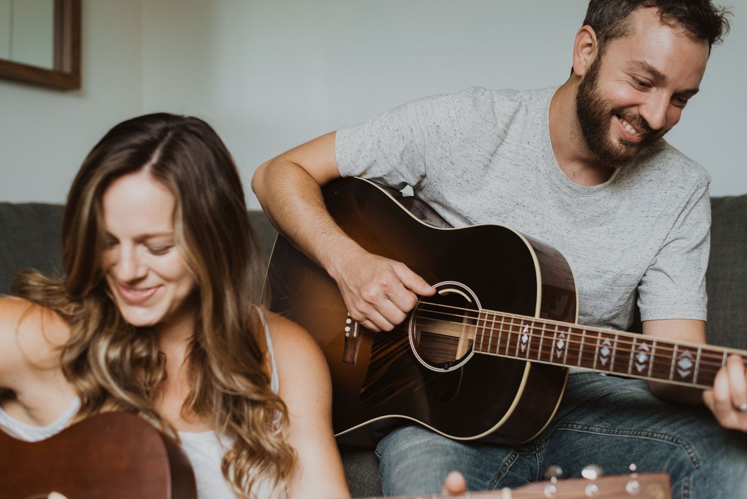 A smiling couple playing acoustic guitars together on a cozy sofa, enjoying a musical moment at home.