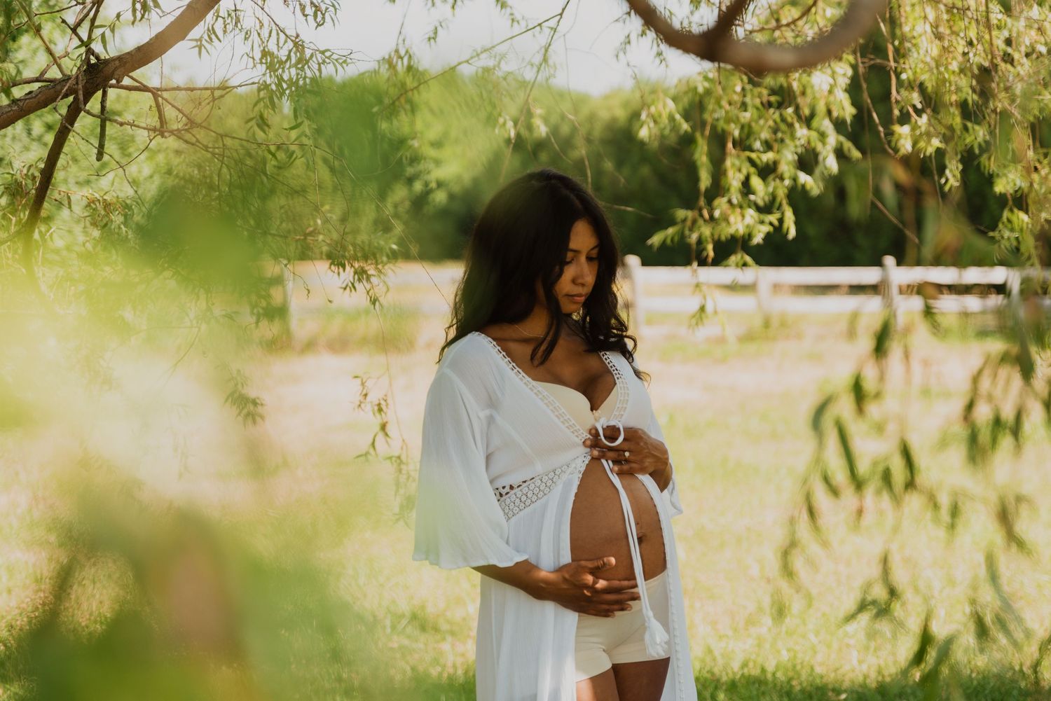 Pregnant woman in white dress cradling baby bump outdoors under green trees during a maternity photo shoot.