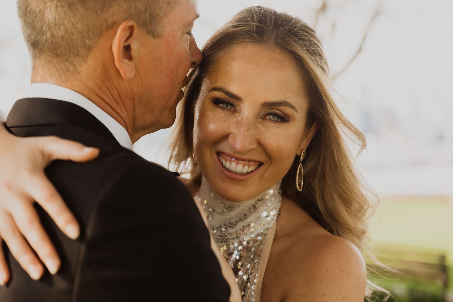 A smiling bride in a sequined dress embraces a man in a tuxedo at an outdoor wedding celebration.