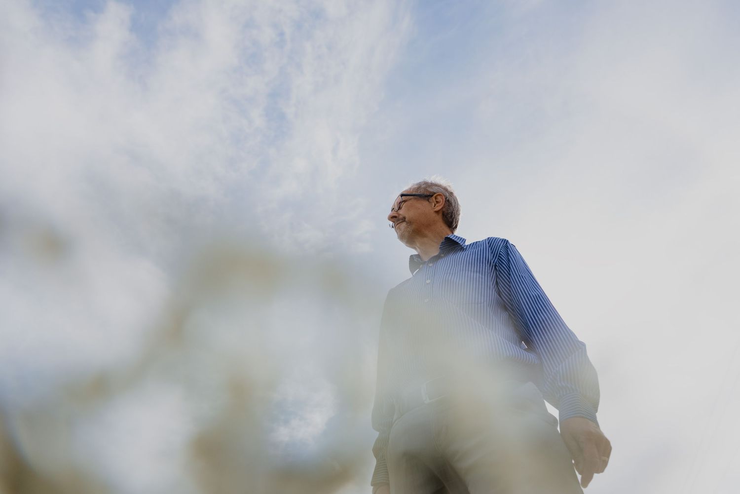 Elderly man in blue jacket gazing upward against a bright cloudy sky, photographed from a low angle.