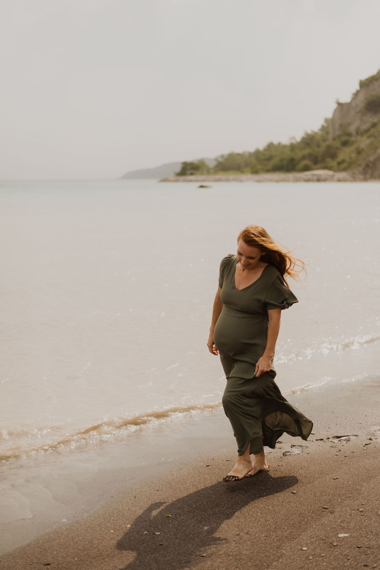 Pregnant woman in olive green dress walking barefoot along a serene beach shoreline with hills in background.