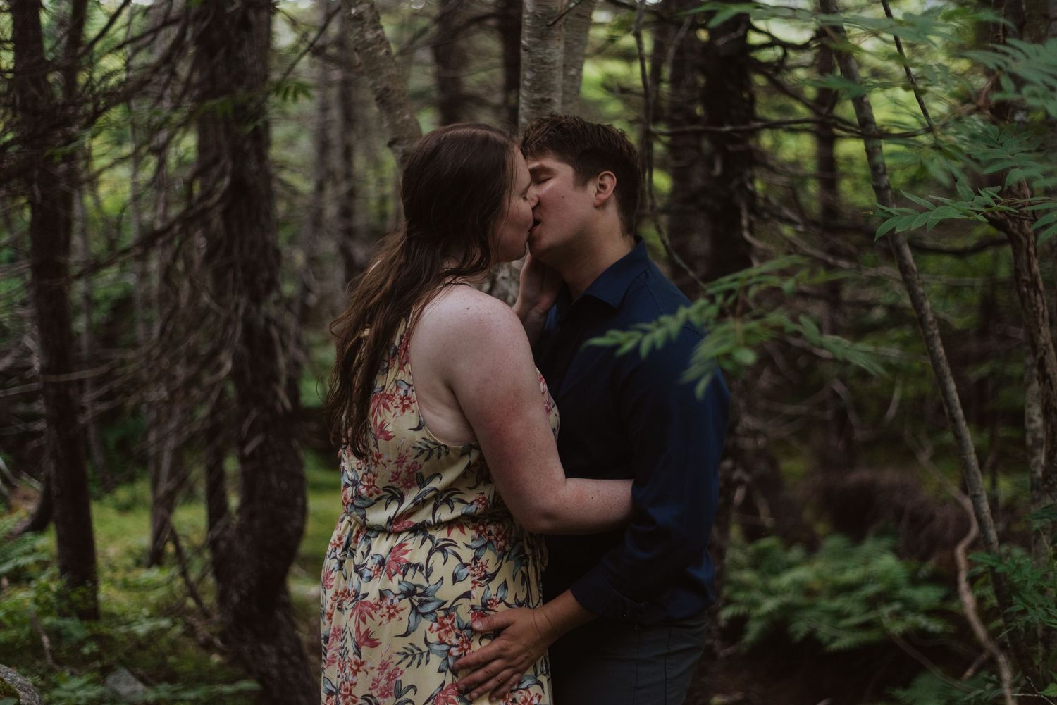 Couple sharing a romantic kiss in a lush forest setting, woman in floral dress embracing partner among green ferns and trees.