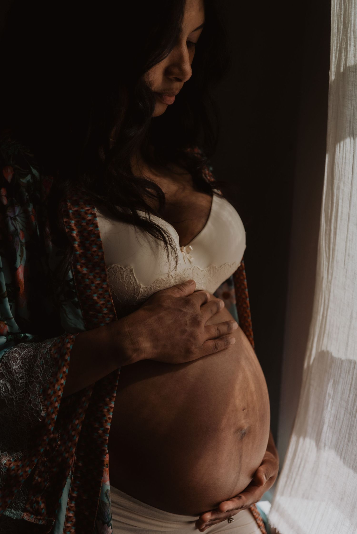 Pregnant woman cradling her baby bump near a window in soft natural light, wearing a white bra and boho kimono.