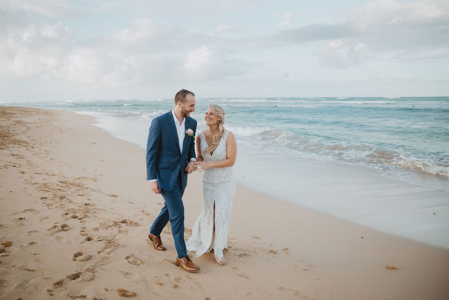 Bride and groom walking hand in hand on a sandy beach with ocean waves, wearing wedding attire on a cloudy day.