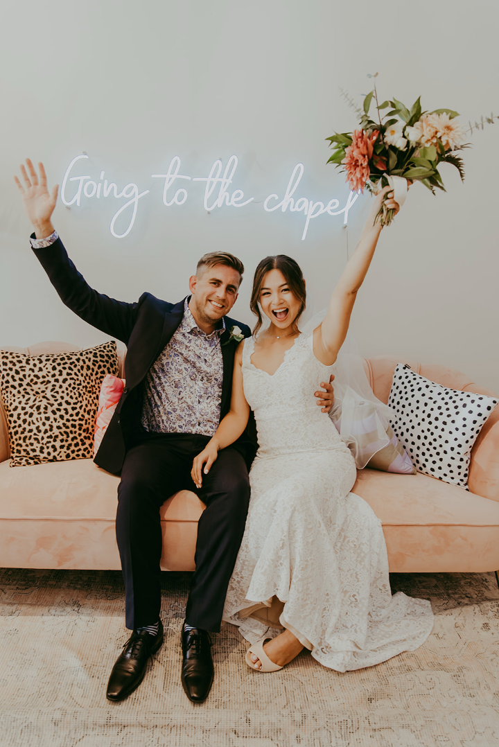 Joyful newlyweds celebrate on a pink sofa beneath a neon Going to the Chapel sign, bride holding bouquet.