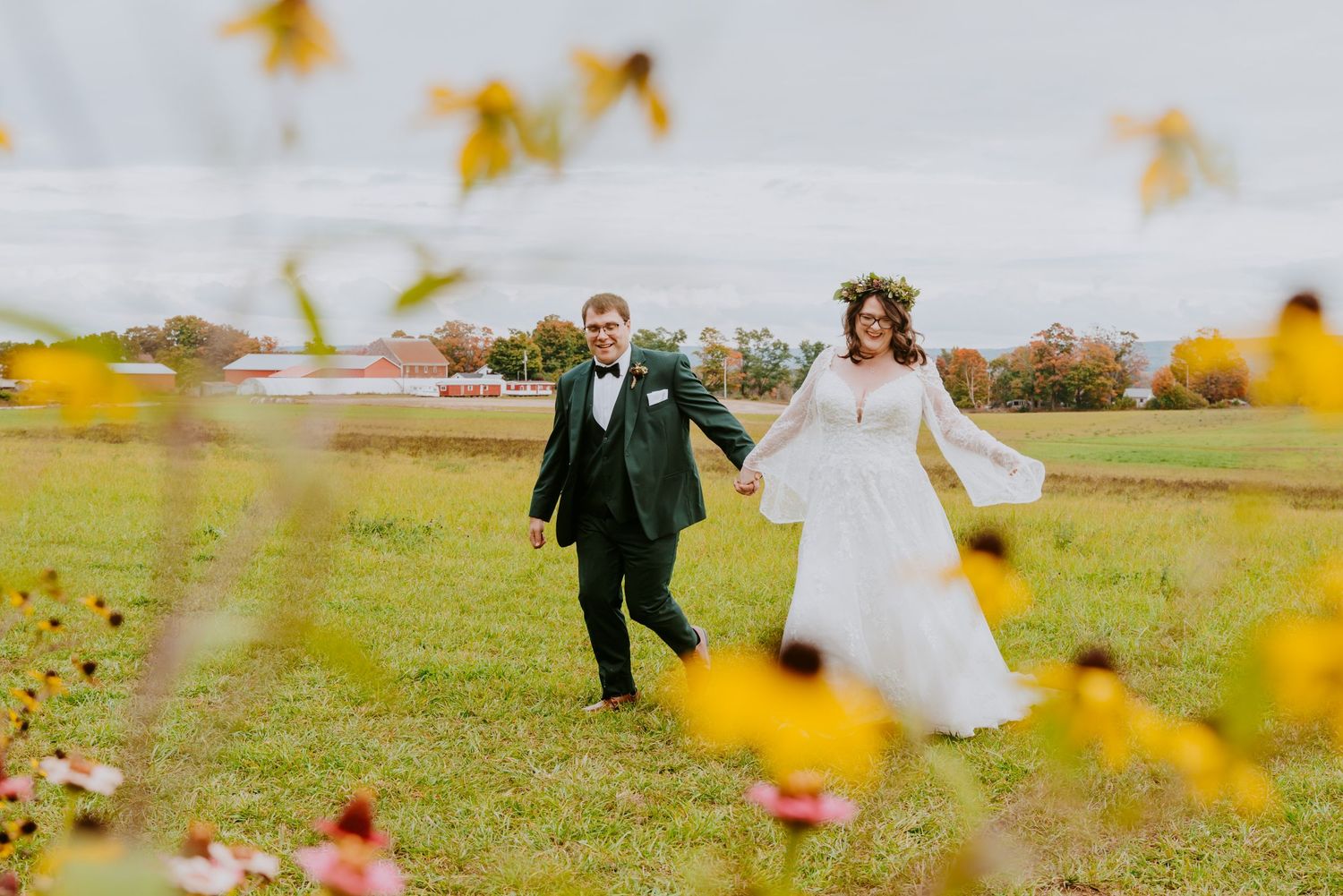 Bride and groom walk hand-in-hand through a green field framed by yellow wildflowers on a cloudy autumn wedding day.