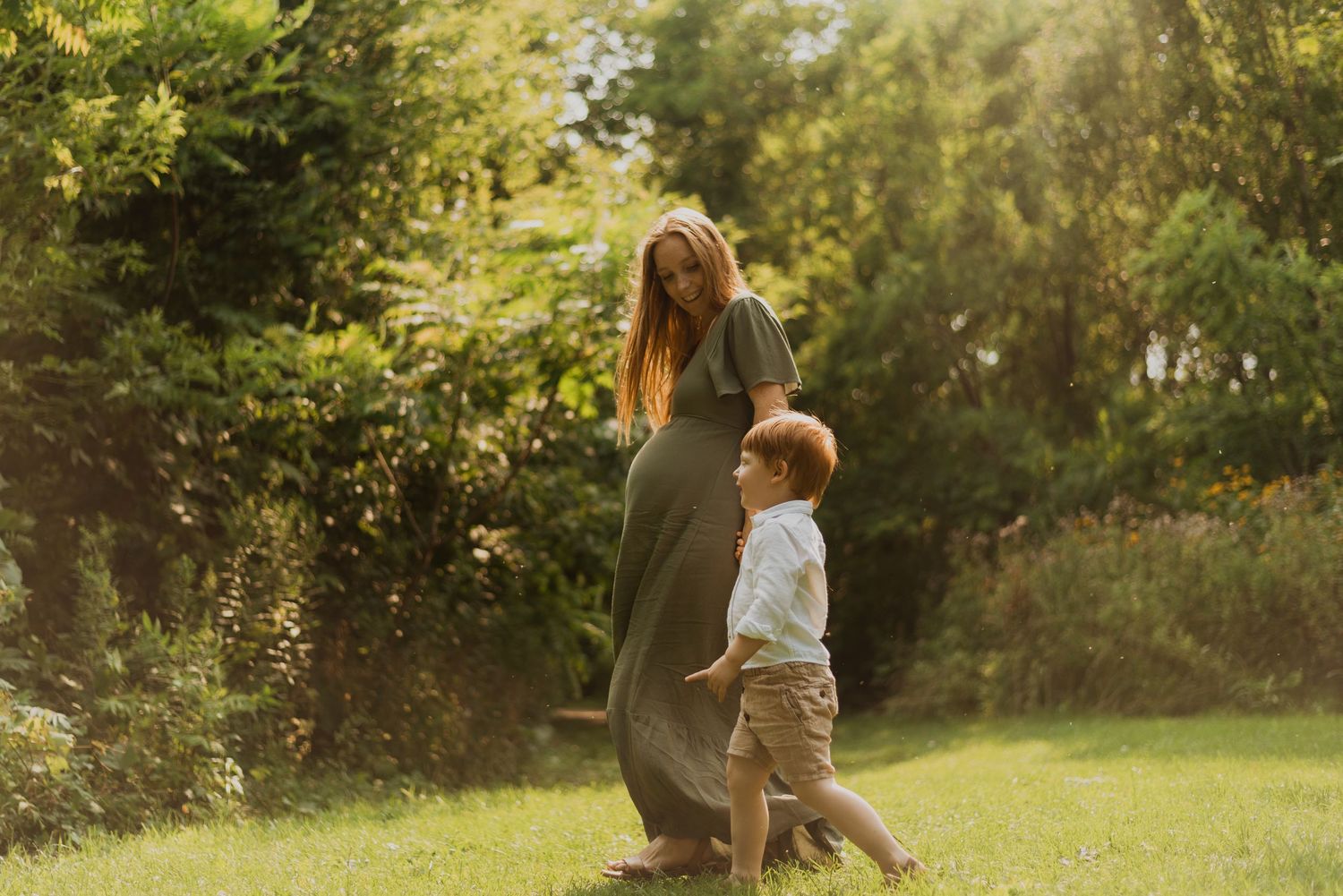 Pregnant woman in green dress walking with toddler boy in sunny garden park setting.