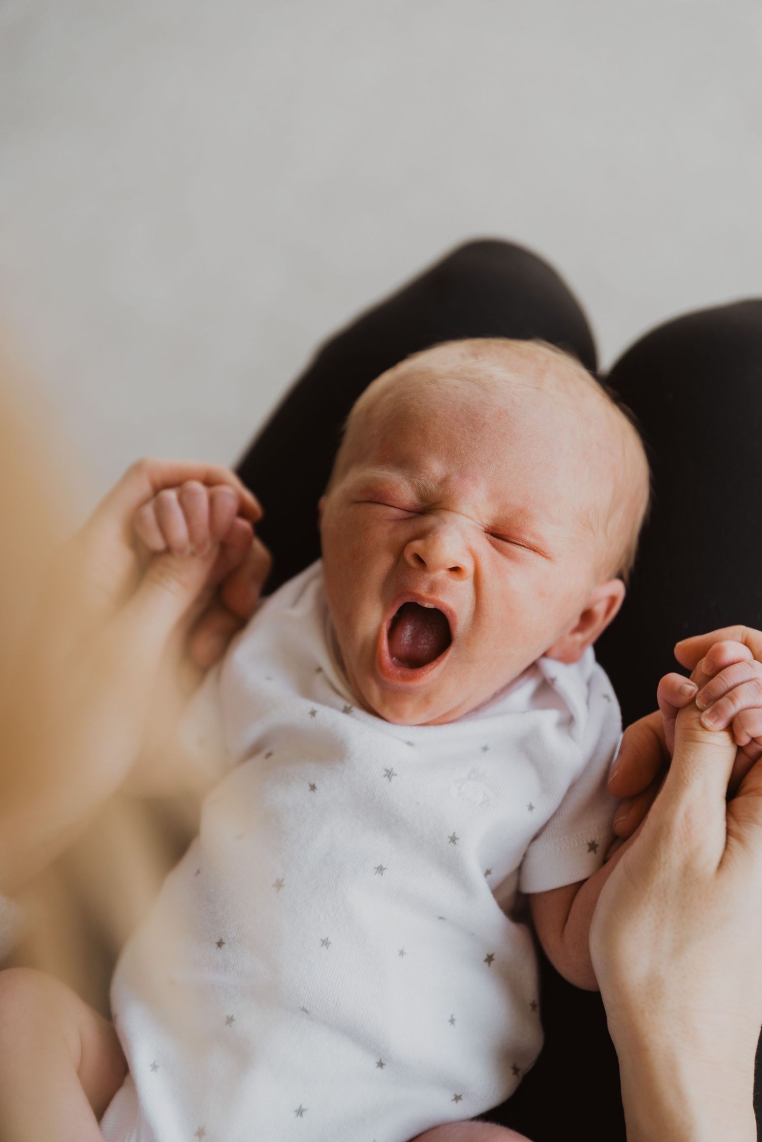 Newborn baby yawning widely while being held by a parent, dressed in a white onesie with small dots.