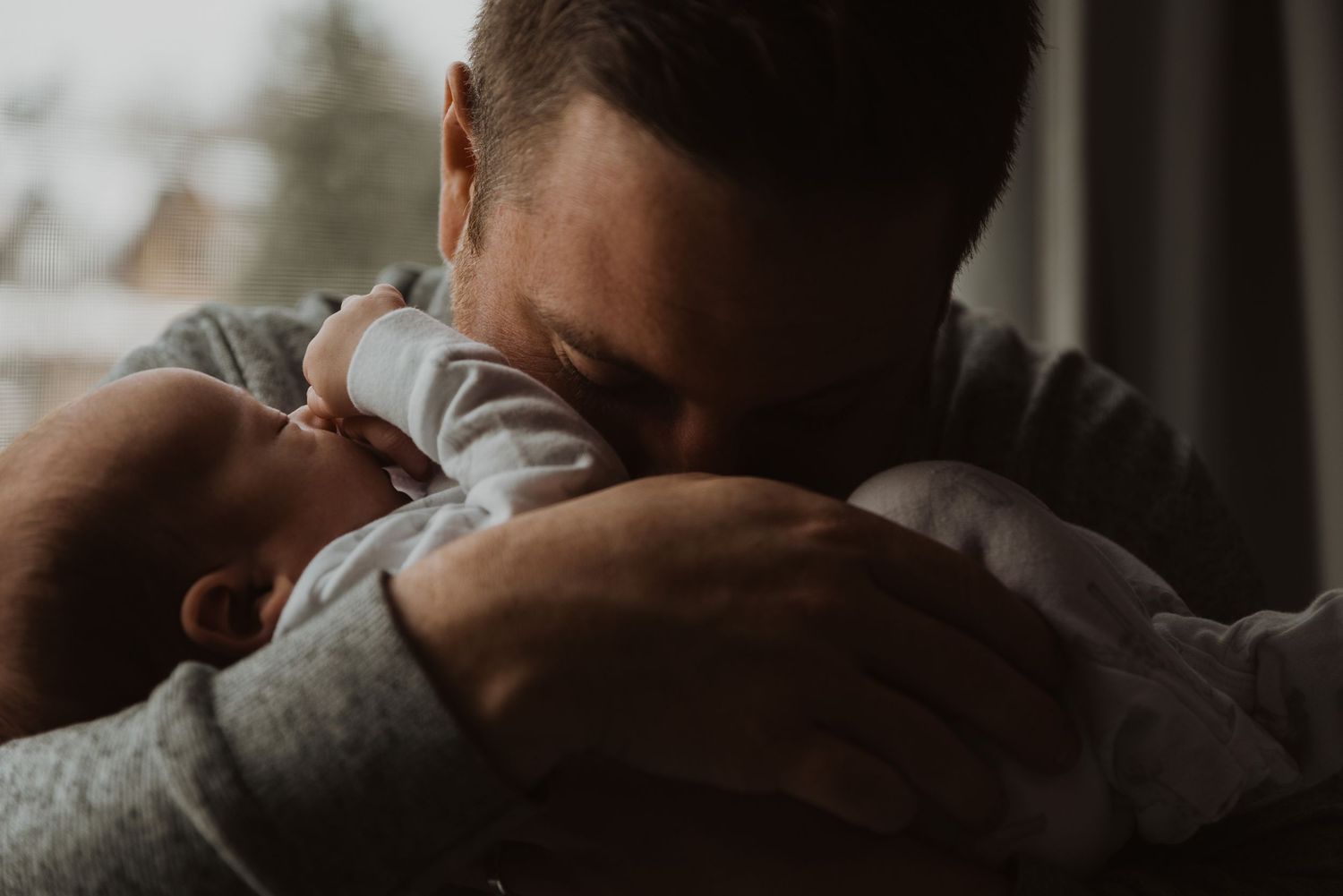 A father tenderly holds and nuzzles his newborn baby, sharing an intimate bonding moment near a window.