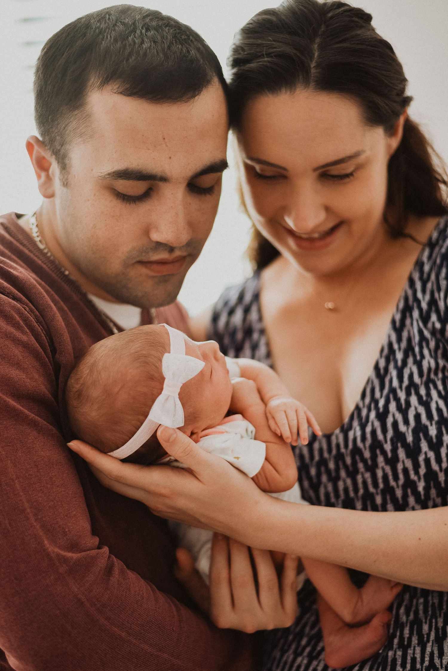 New parents lovingly cradling their sleeping newborn baby girl wearing a white bow headband.