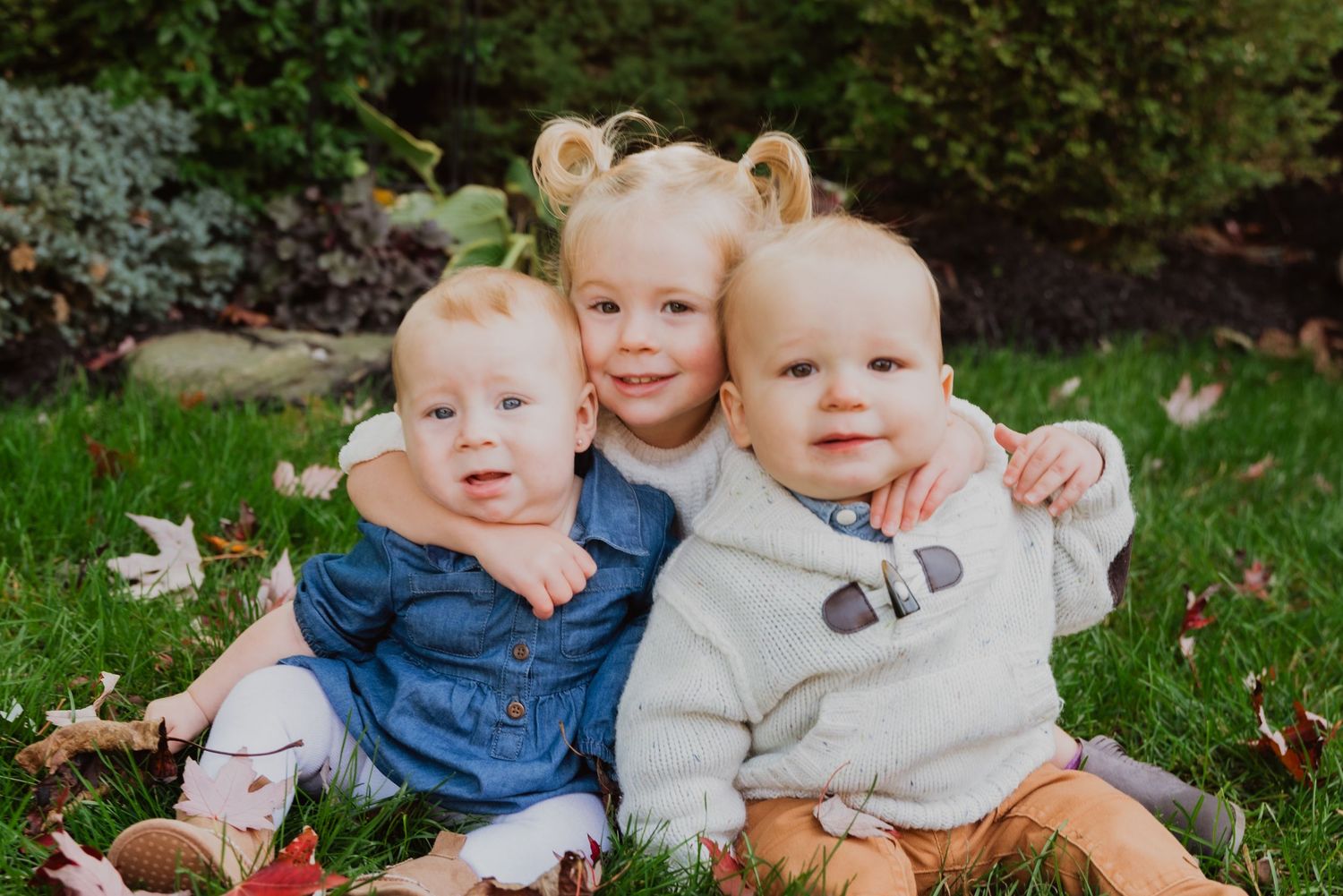 Three young siblings sit together on green grass surrounded by autumn leaves, smiling warmly in a charming outdoor portrait.