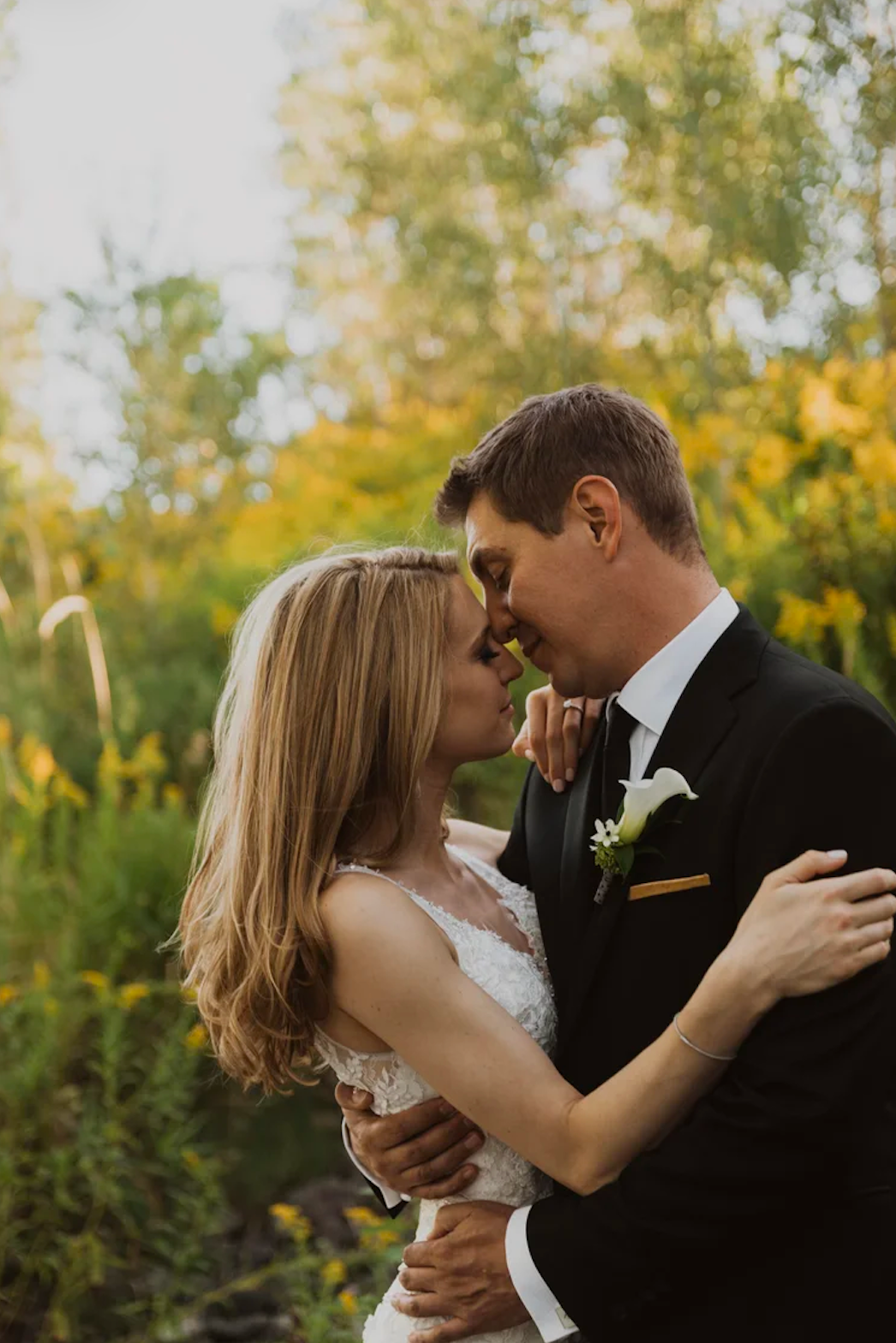 A bride and groom share a romantic kiss outdoors, surrounded by lush yellow and green foliage on their wedding day.