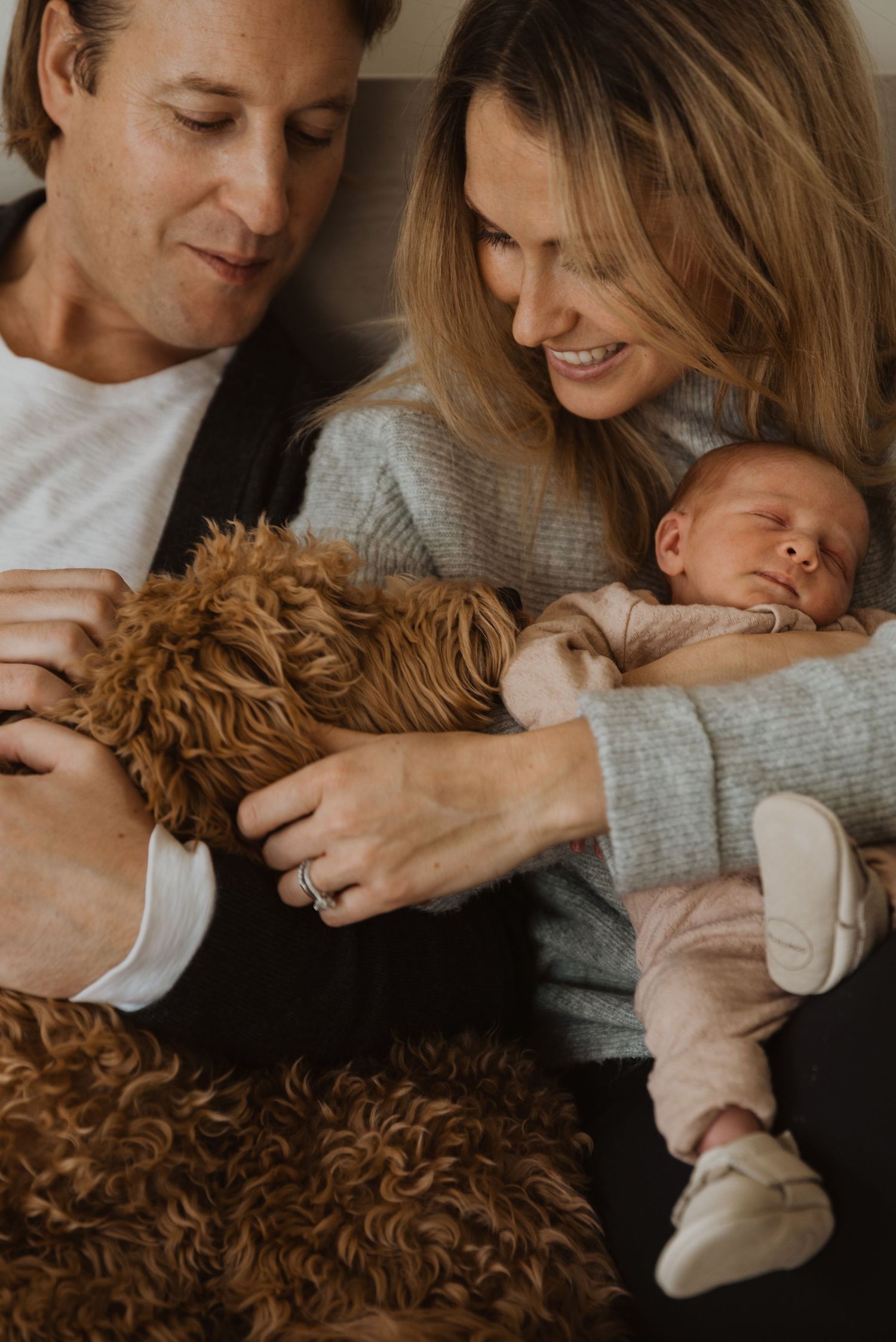 A couple smiles at their sleeping newborn baby while cuddling a fluffy brown dog on a cozy sofa.