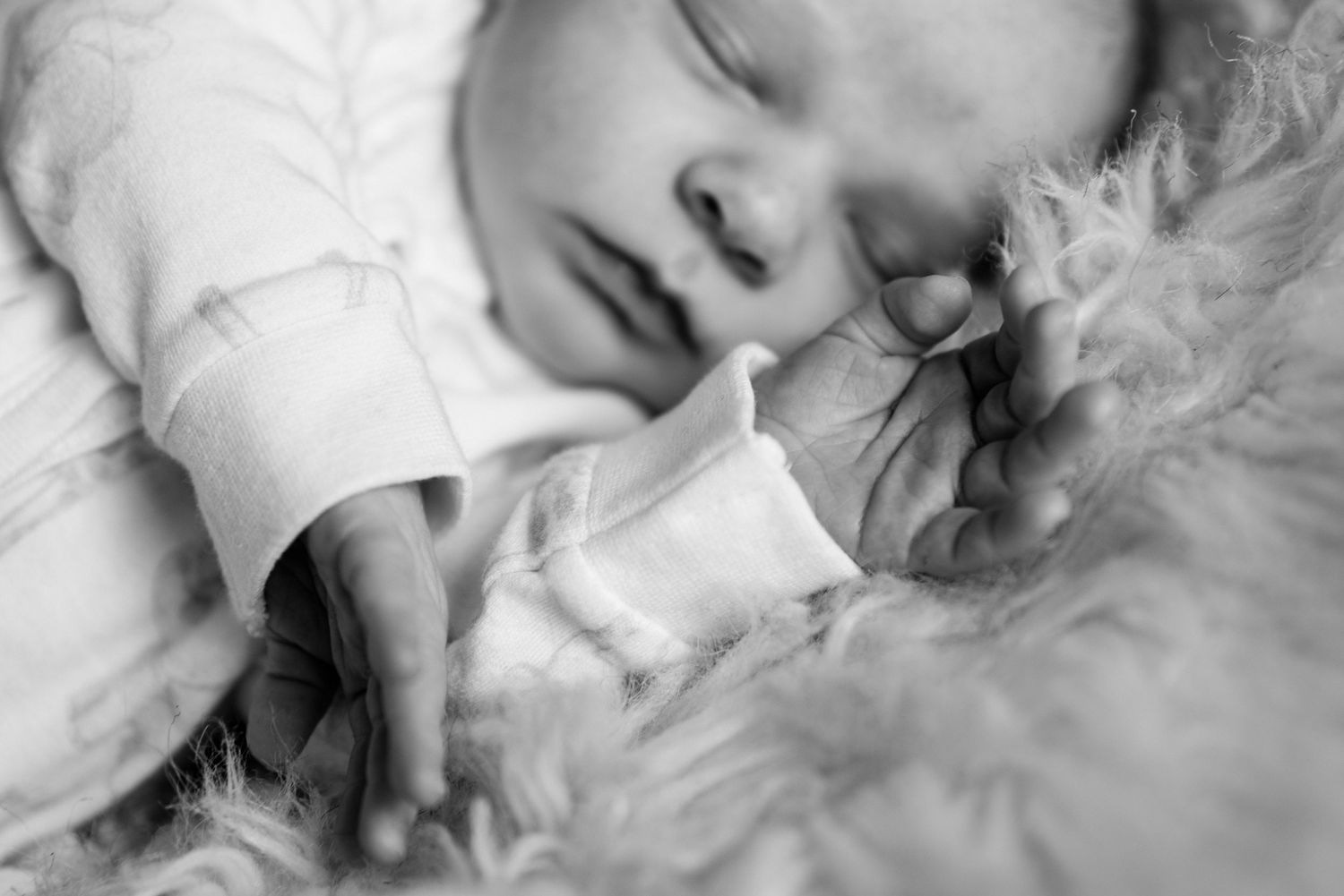 Black and white photo of a sleeping newborn baby in a soft fur blanket, wearing a white onesie.