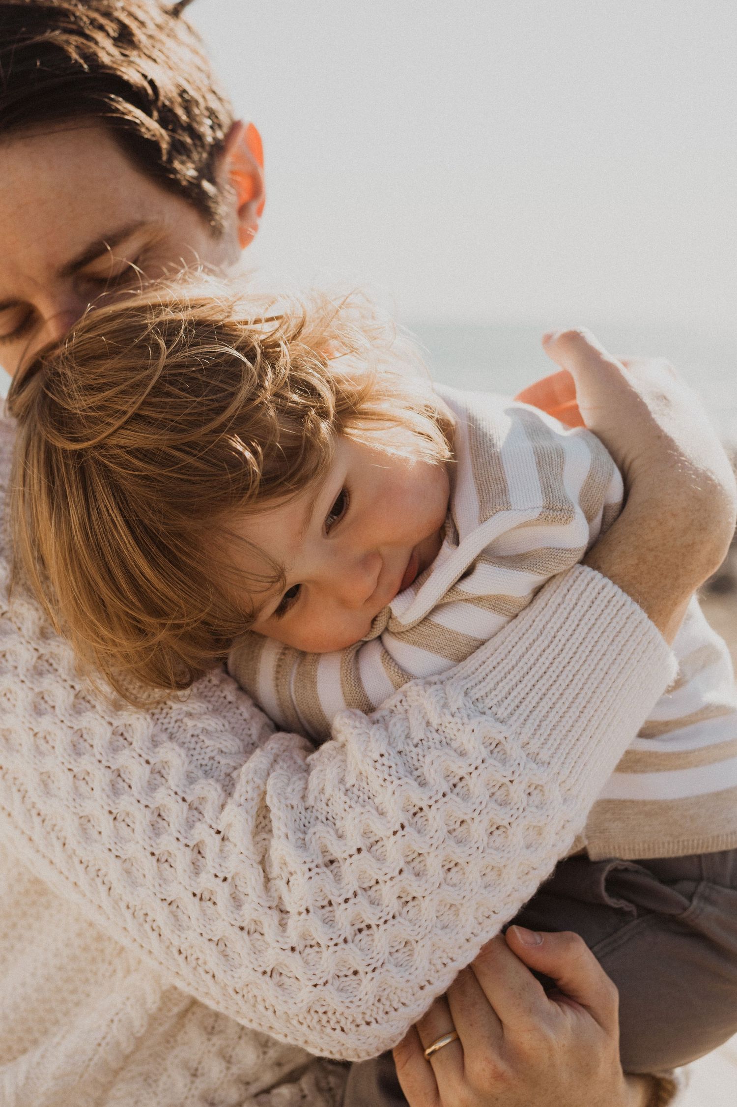 A parent in a white knit sweater tenderly holds a young child in a striped shirt outdoors.
