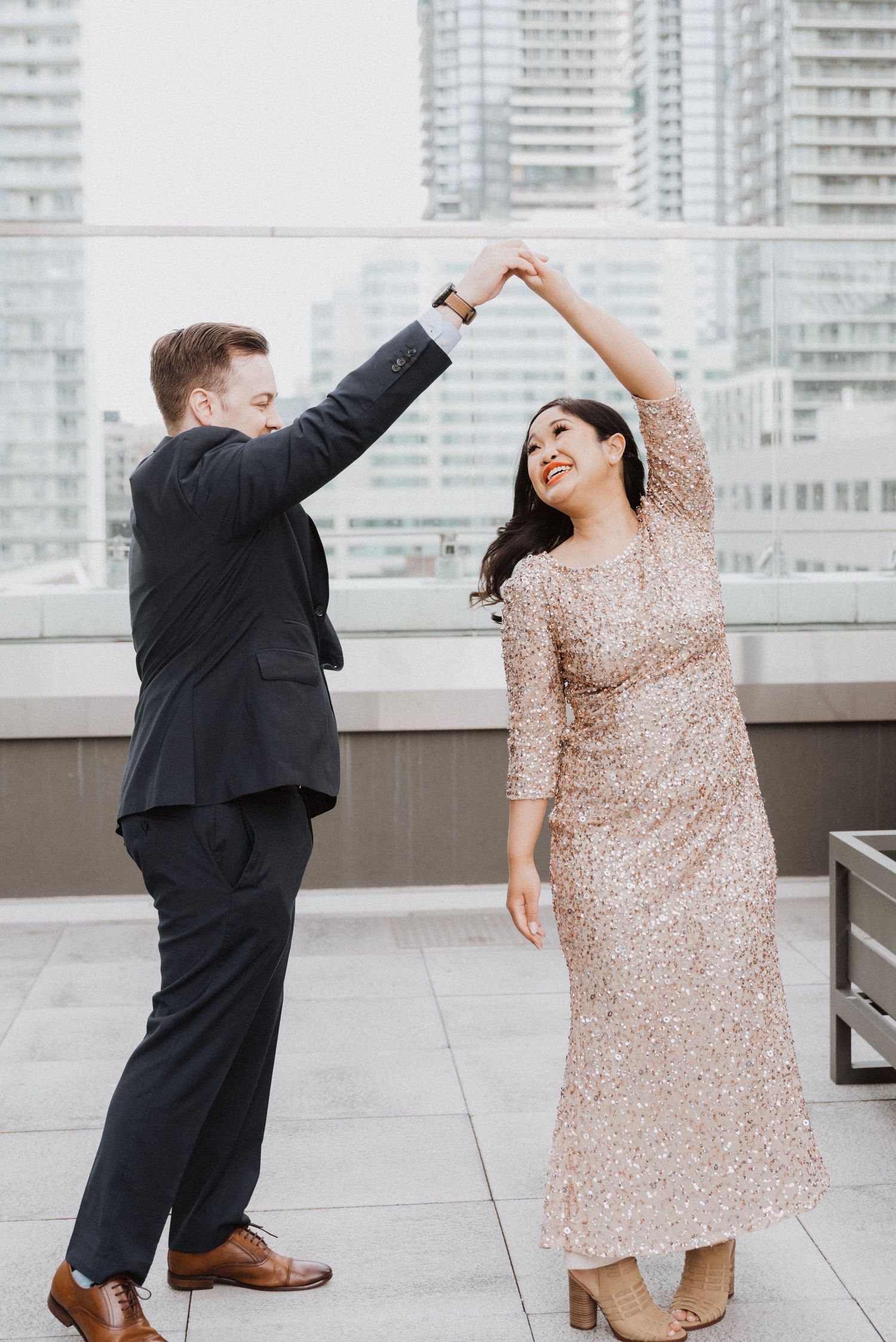 A couple dances on a rooftop terrace, man in black suit and woman in gold sequin dress, with city skyline backdrop.