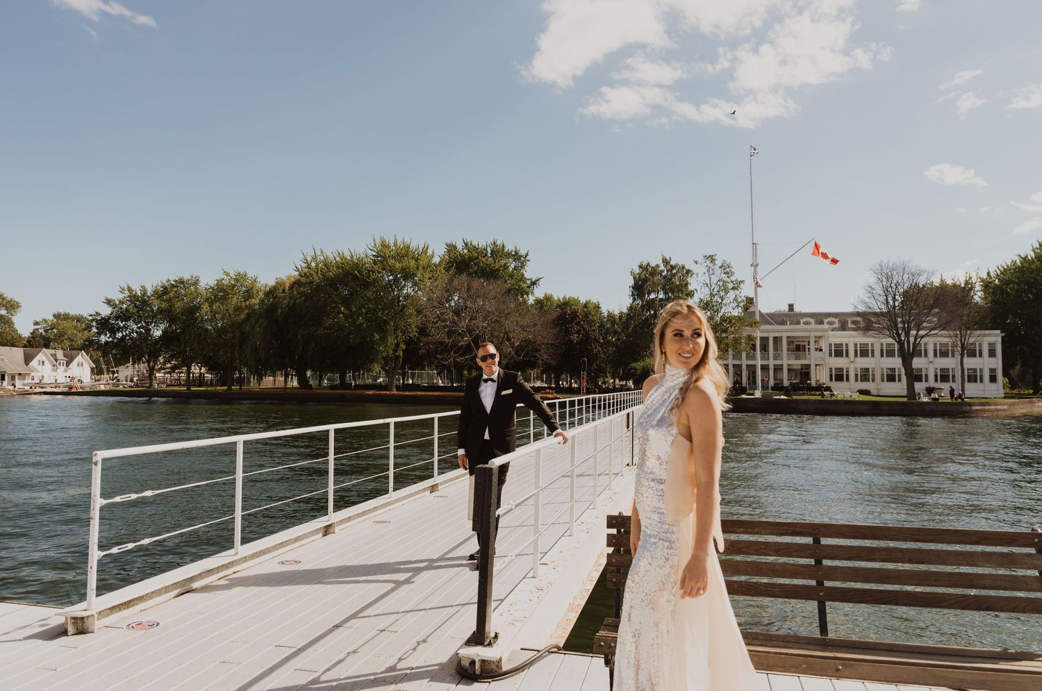 Bride and groom posing on a waterfront dock with a Canadian flag and white building in the background on a sunny day.