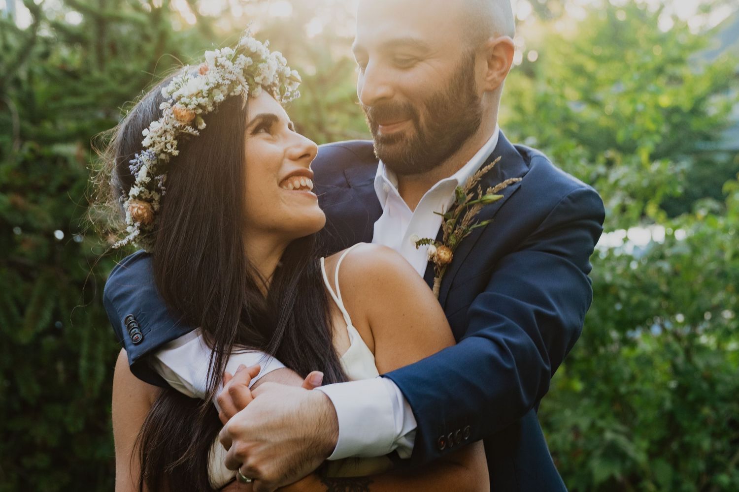 A smiling bride wearing a floral crown embraces her groom in a navy suit outdoors in golden sunlight.