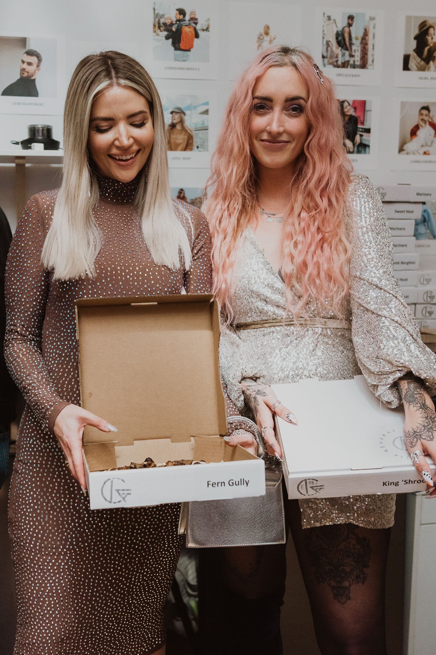 Two women in elegant dresses holding open gift boxes labeled Fern Gully and King Sh at an indoor event.