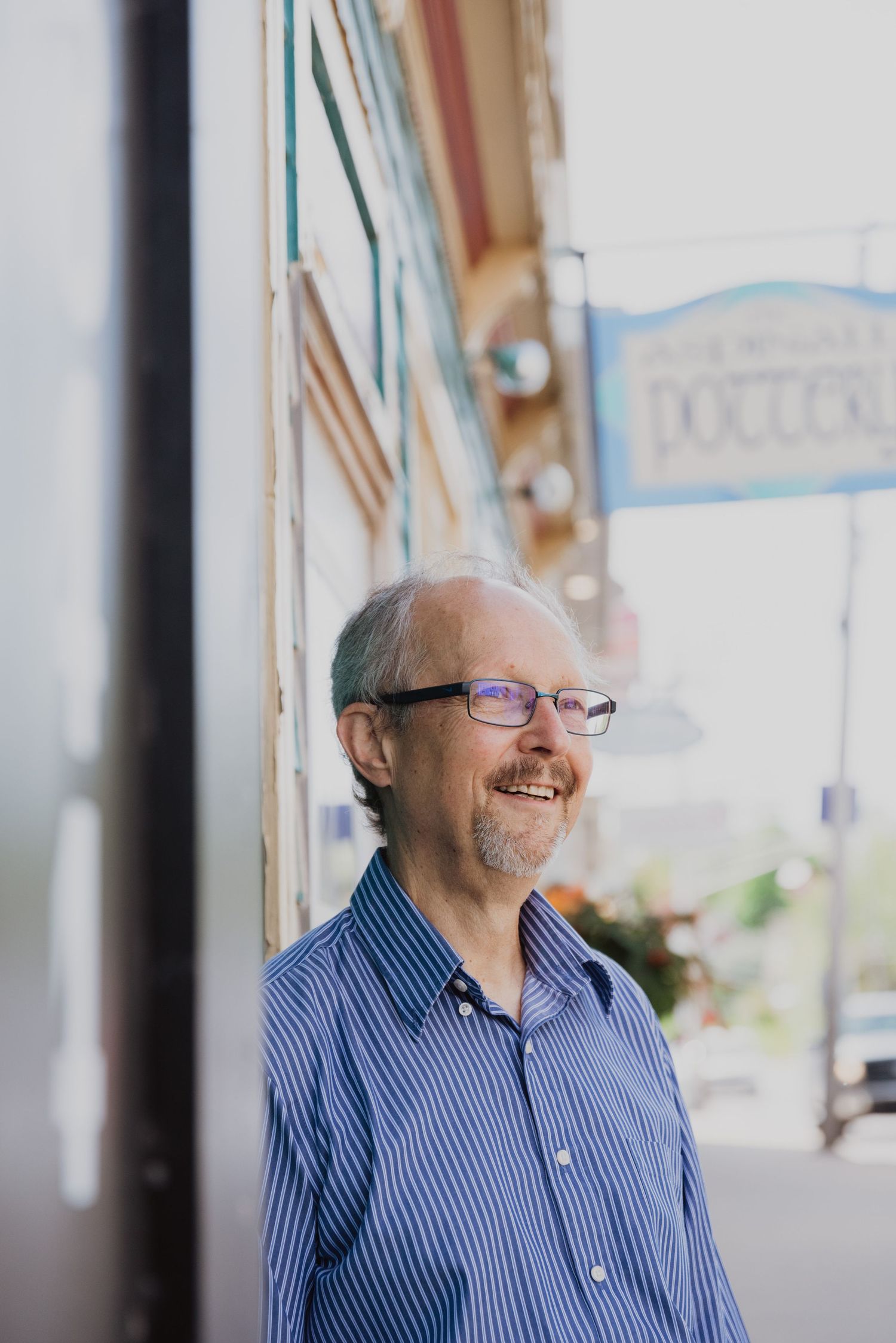 Smiling middle-aged man with glasses and blue striped shirt standing outside a storefront on a sunny day.