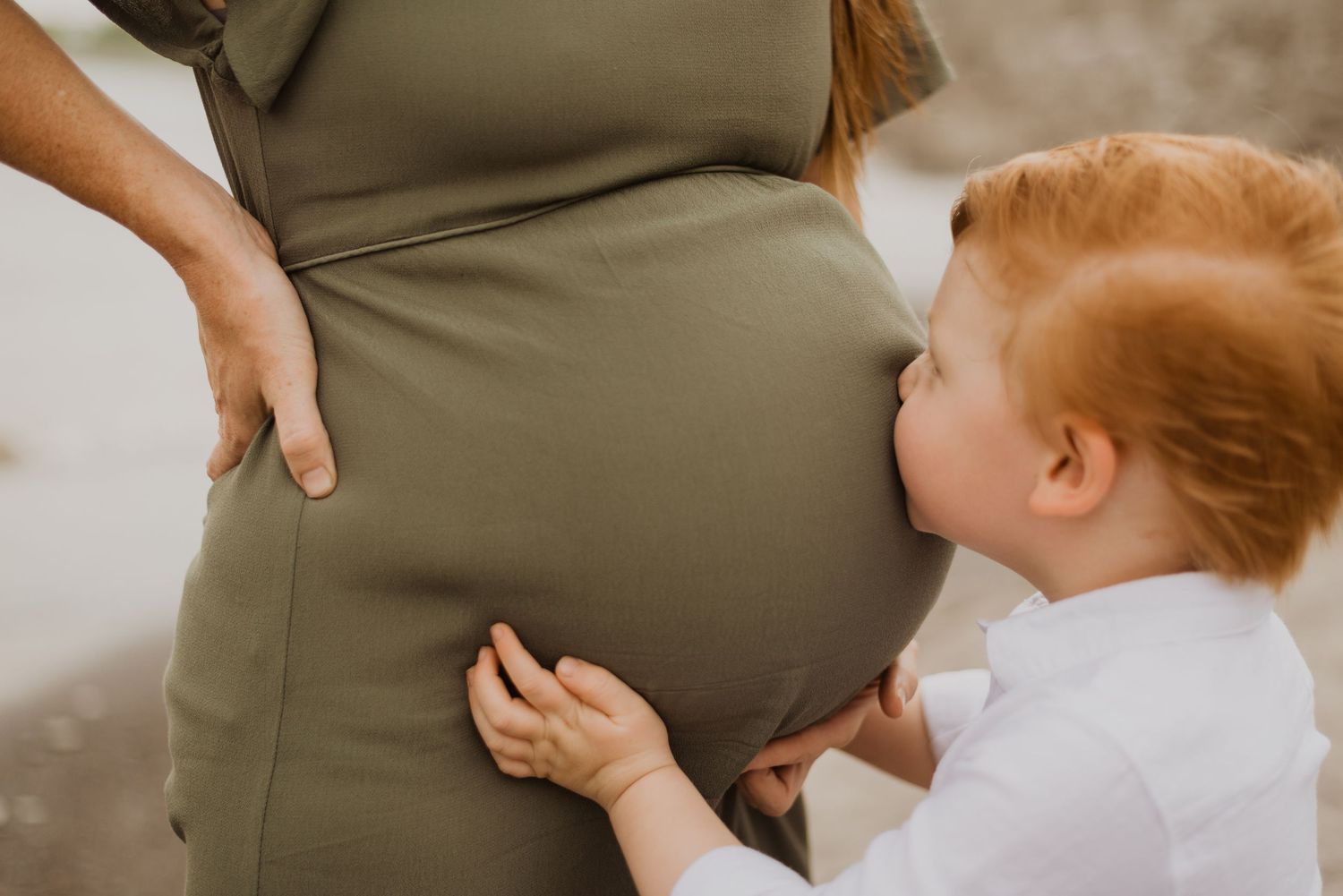 A red-haired toddler kissing his pregnant mother's baby bump while she wears an olive green maternity dress.