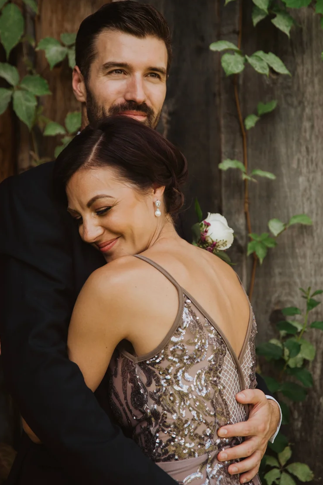 Couple embracing outdoors, woman in sequined dress with white flower, man in black suit, against rustic wood wall.